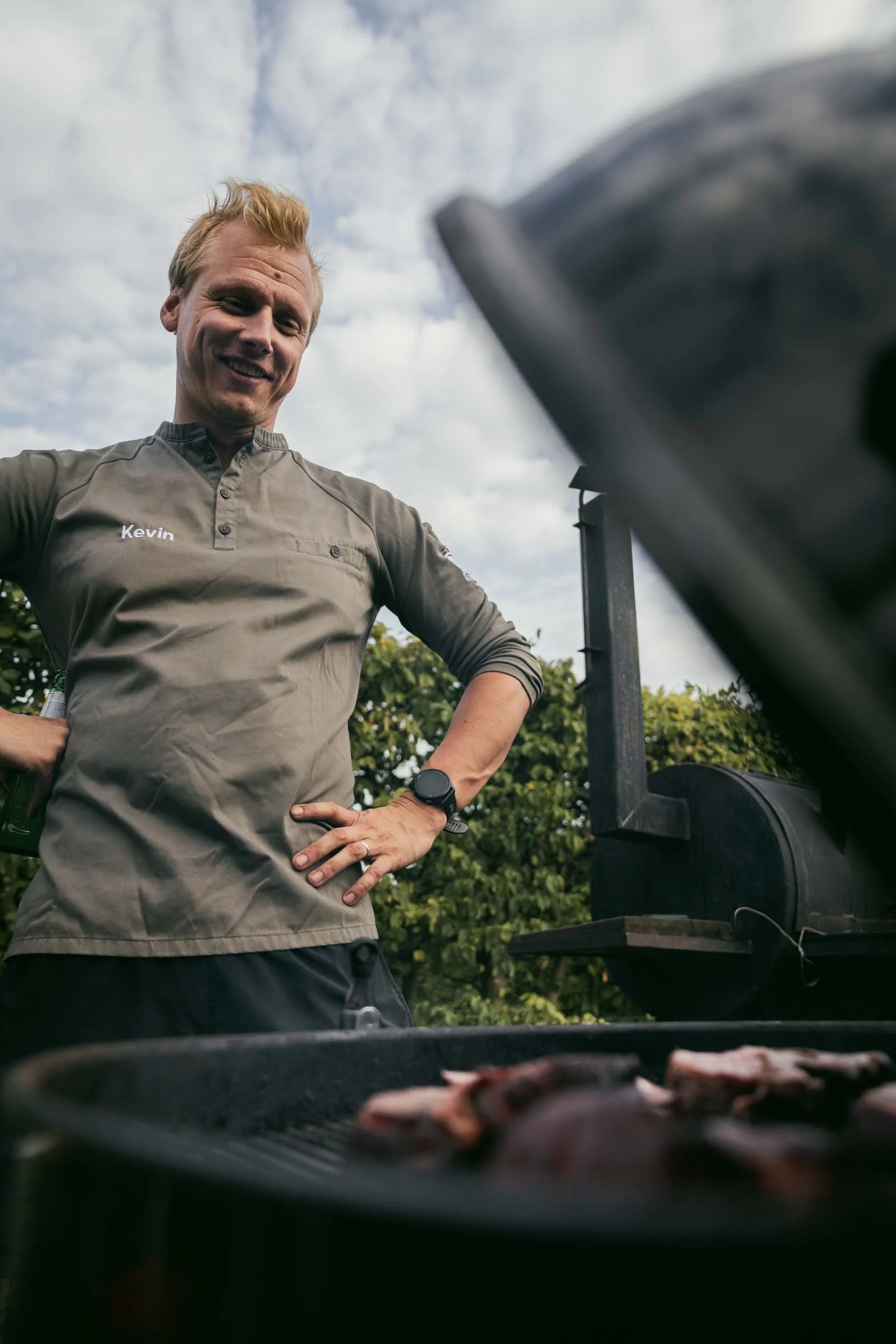 Man with blonde hair smiling, wearing a gray shirt with "Kevin" embroidered on it, standing outdoors near a barbecue grill with meat, under a partly cloudy sky.