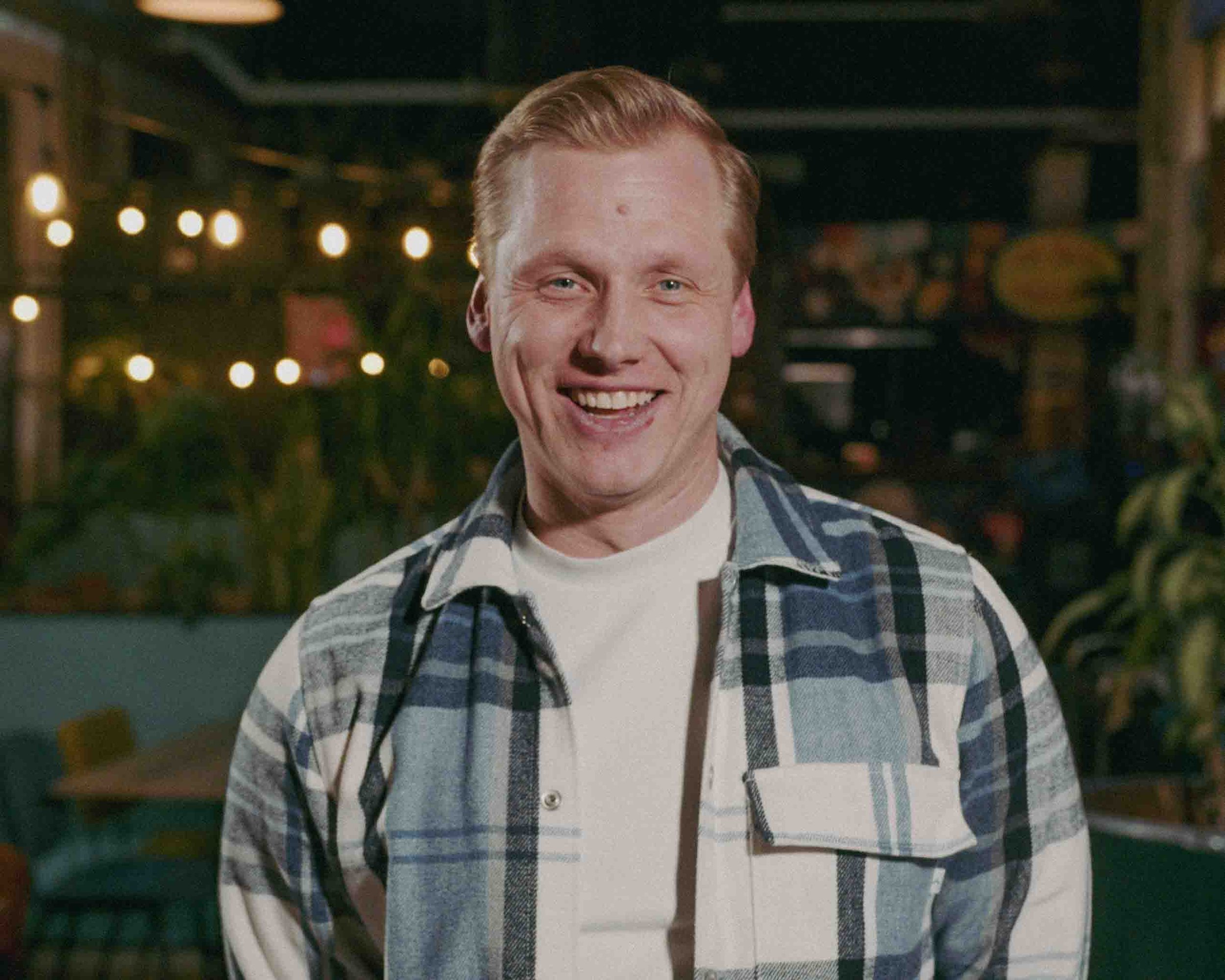 A man in outdoor cooking attire stands with hands on hips, smiling while looking at a barbecue grill with meat, outdoors with green trees and cloudy sky background.
