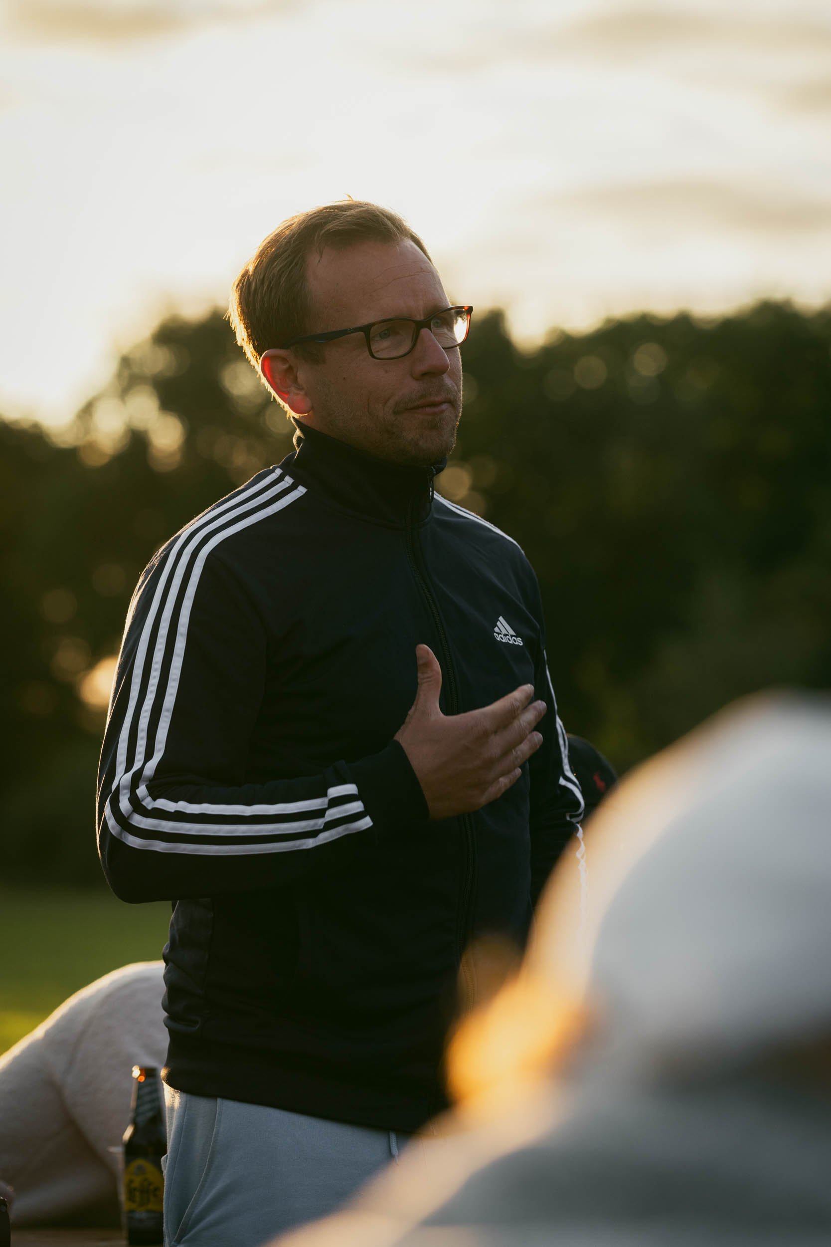 Man wearing glasses and a black Adidas jacket with white stripes, standing outdoors at sunset, with hand on chest, possibly speaking or making a point.
