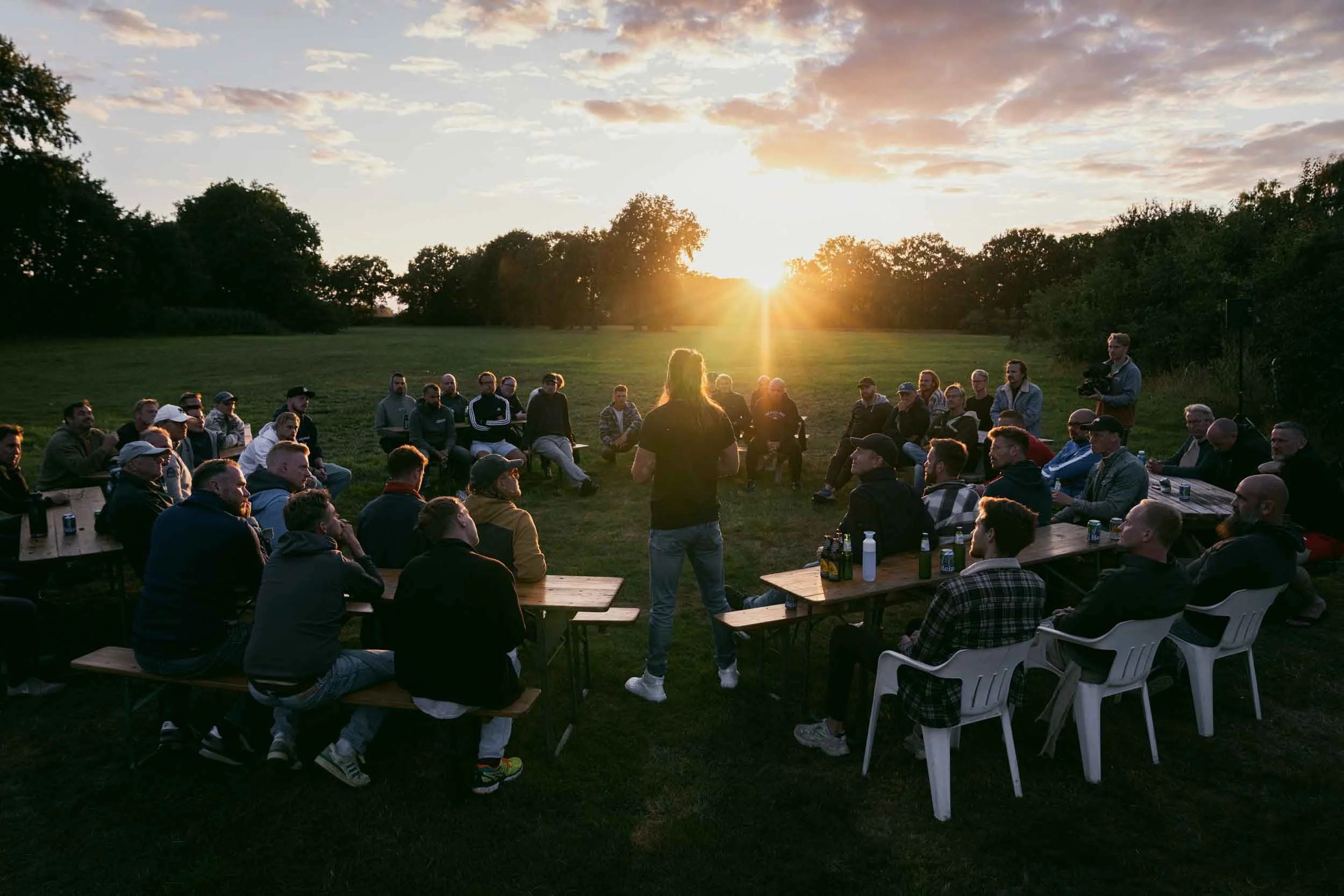 A group of people gathered outdoors in a circle, listening to a speaker at sunset, with trees and an open field in the background.
