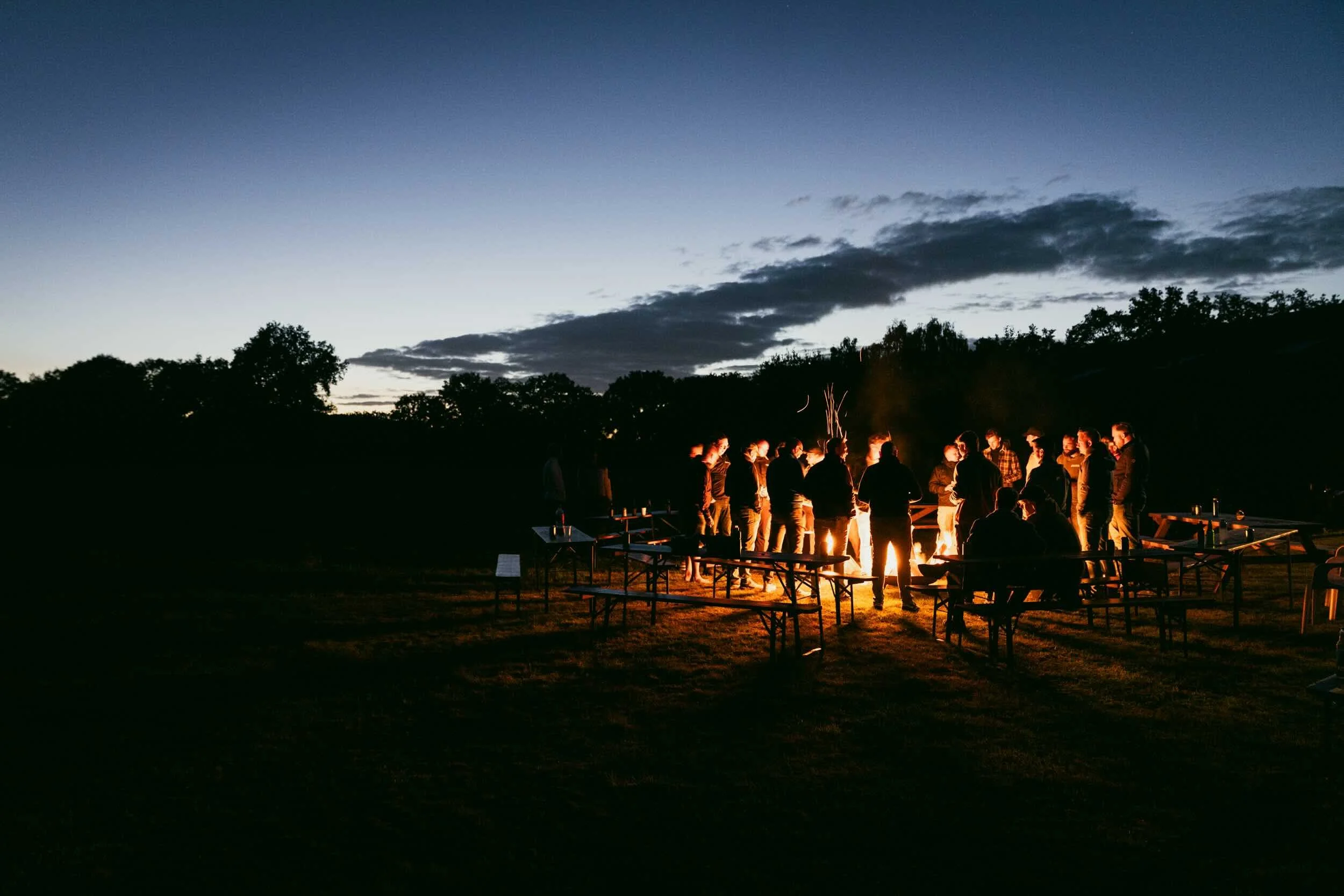 Group of people gathered around a campfire during dusk in an outdoor setting with picnic tables and trees in the background.