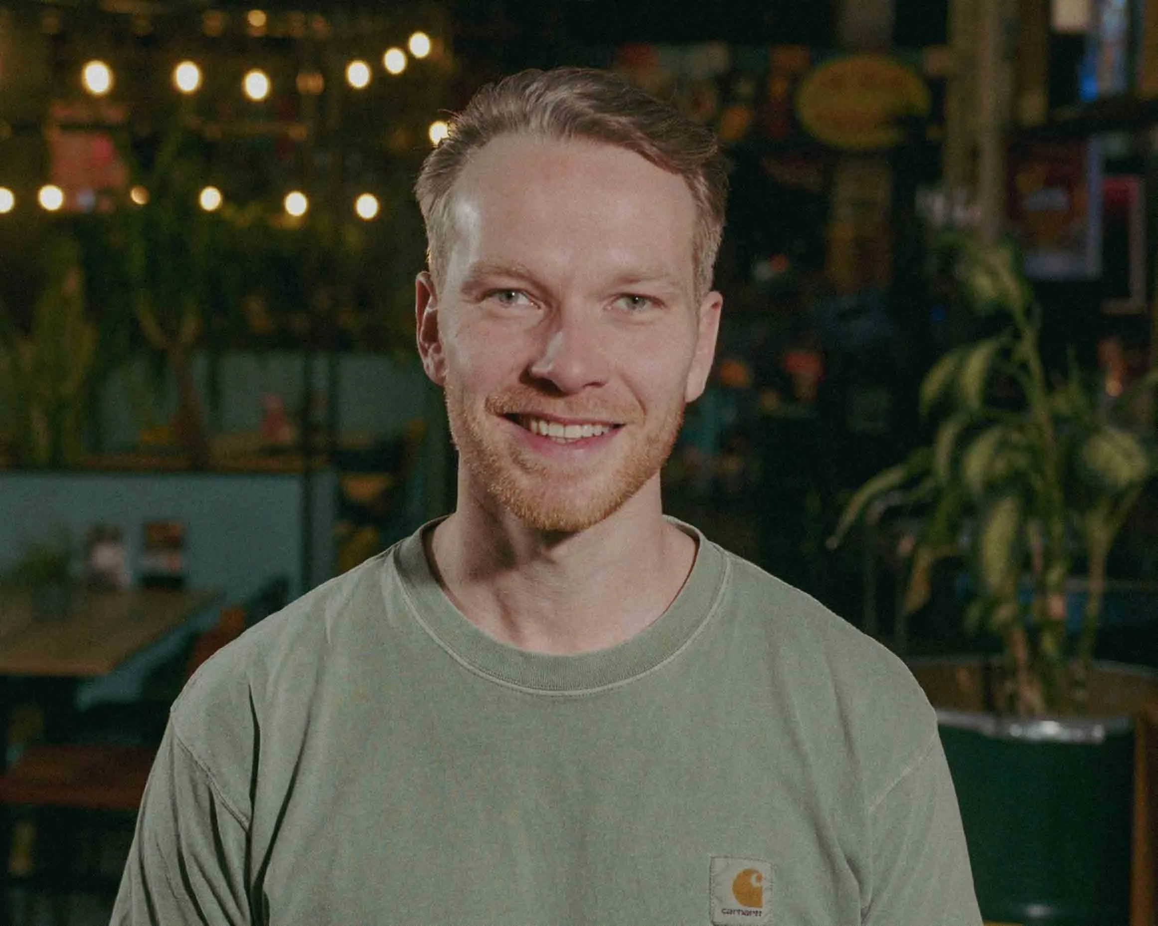 A smiling man with light skin and short brown hair, wearing a black jacket over a white shirt, looking to his left.