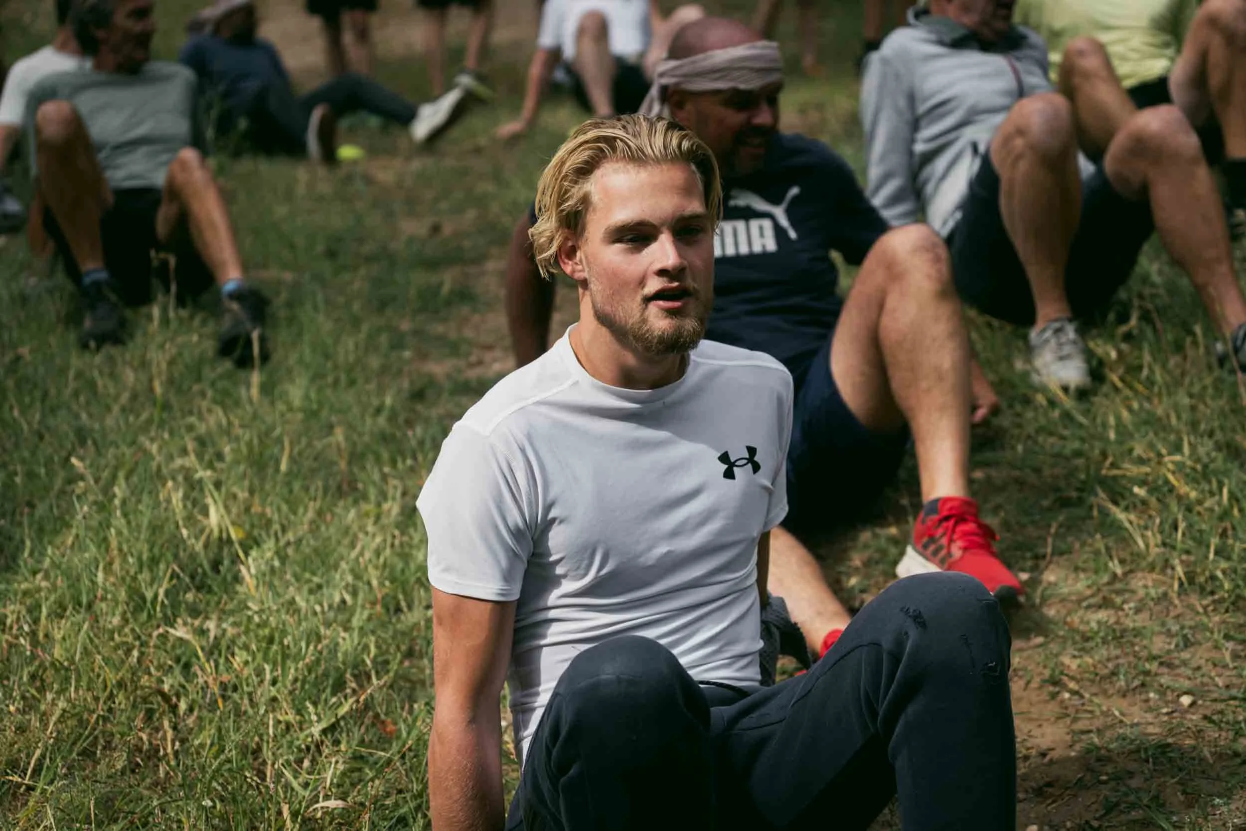 A group of people outdoors on grass, sitting and leaning back, relaxing after exercise. The focus is on a young man with blond hair and a beard wearing a white Under Armour t-shirt.