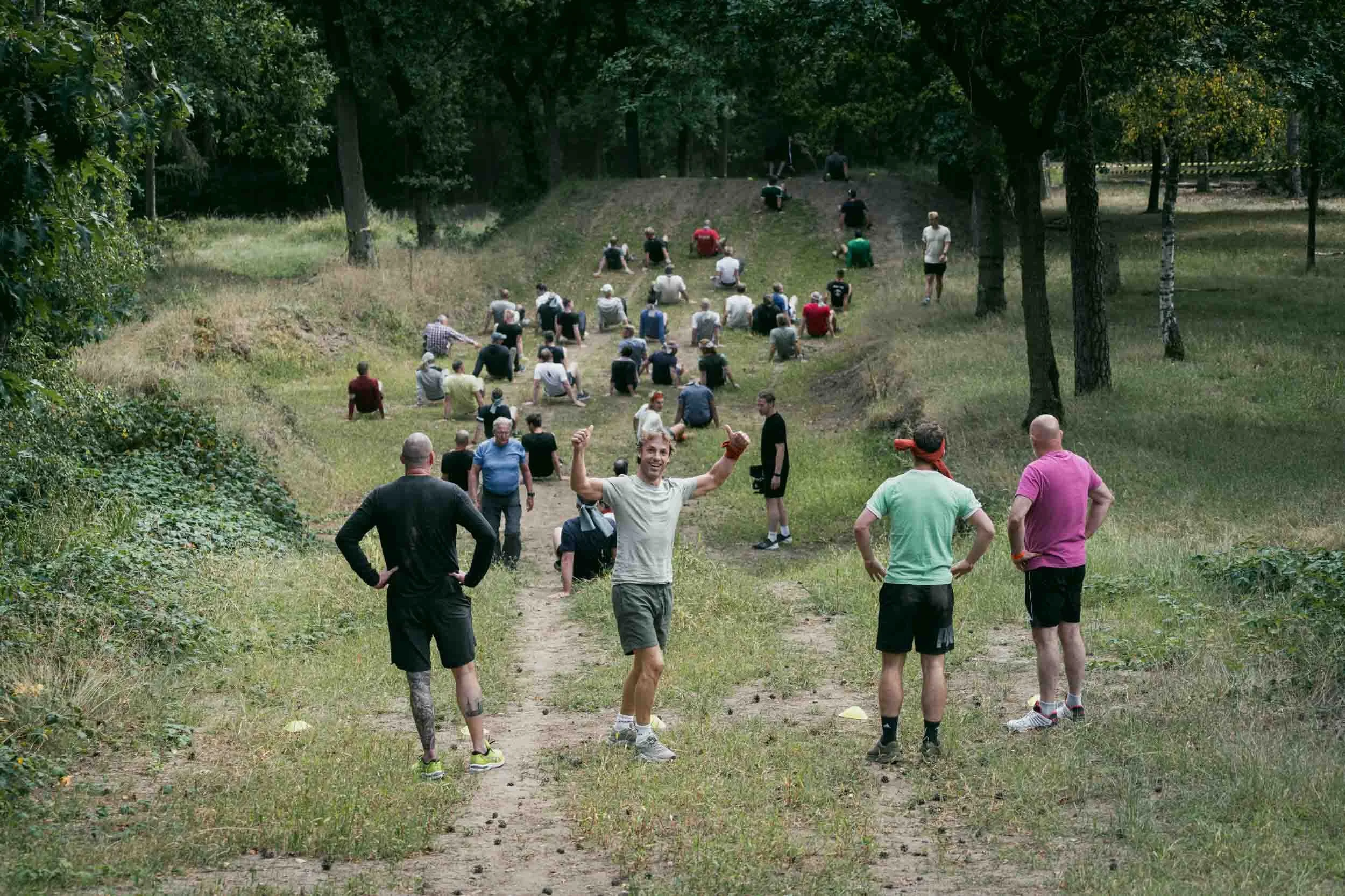 People participating in an outdoor fitness event in a park, some sitting on a slope, others standing on a dirt path surrounded by trees.