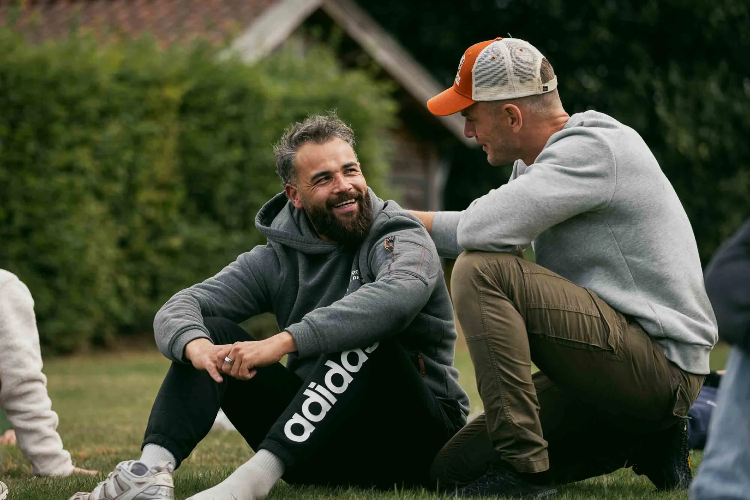 Two men outdoors, one sitting on the grass with a smile, the other kneeling, engaging in conversation, surrounded by greenery.