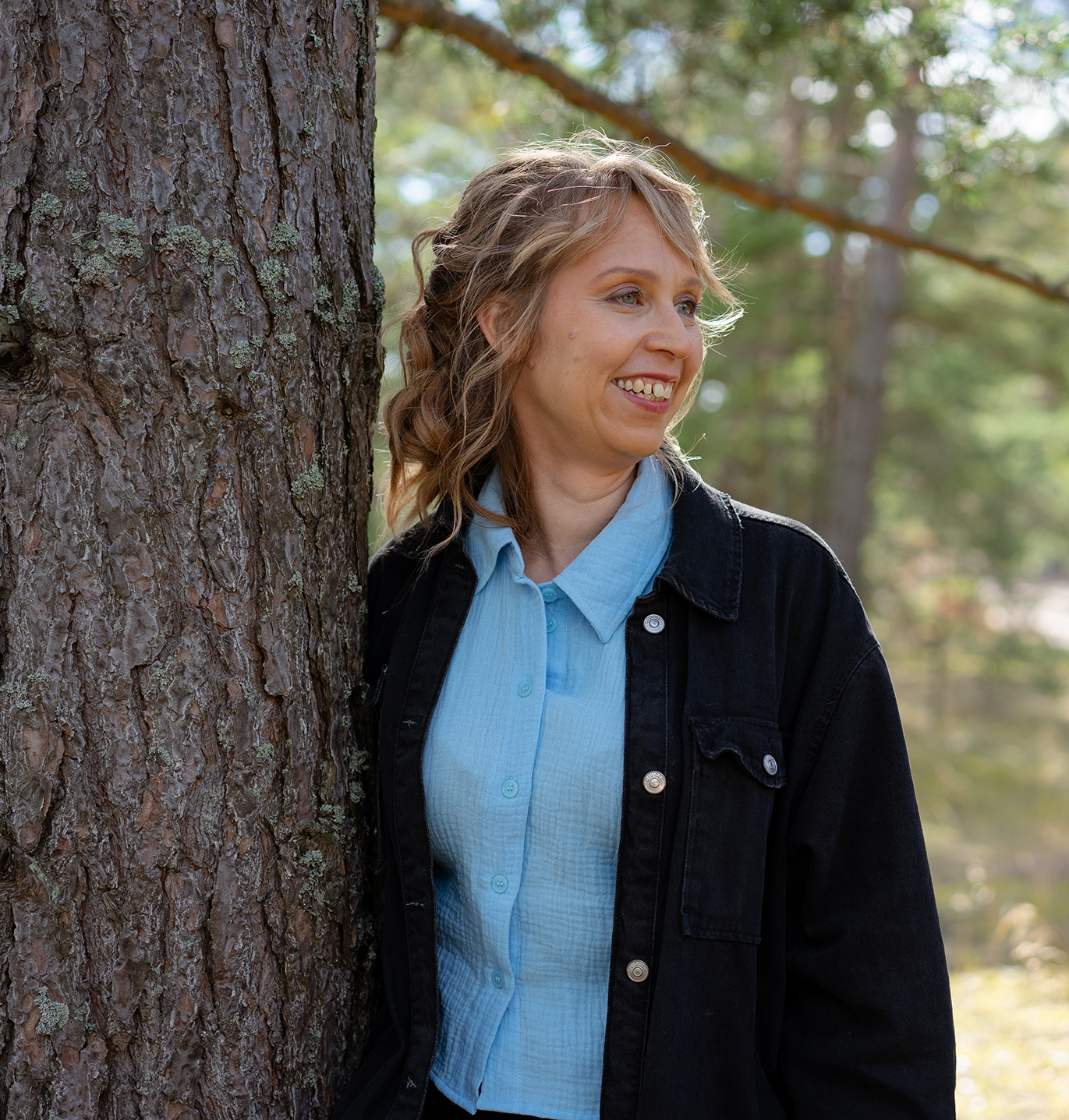 Woman with blonde curly hair smiling and leaning against a tree in a forest.