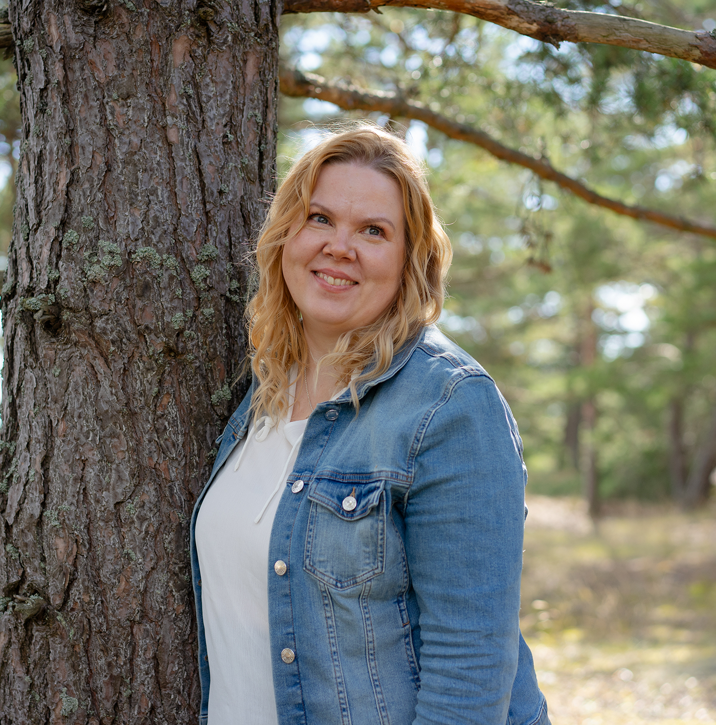 A smiling woman with blonde hair wearing a denim jacket, standing outdoors next to a large tree in a forest.