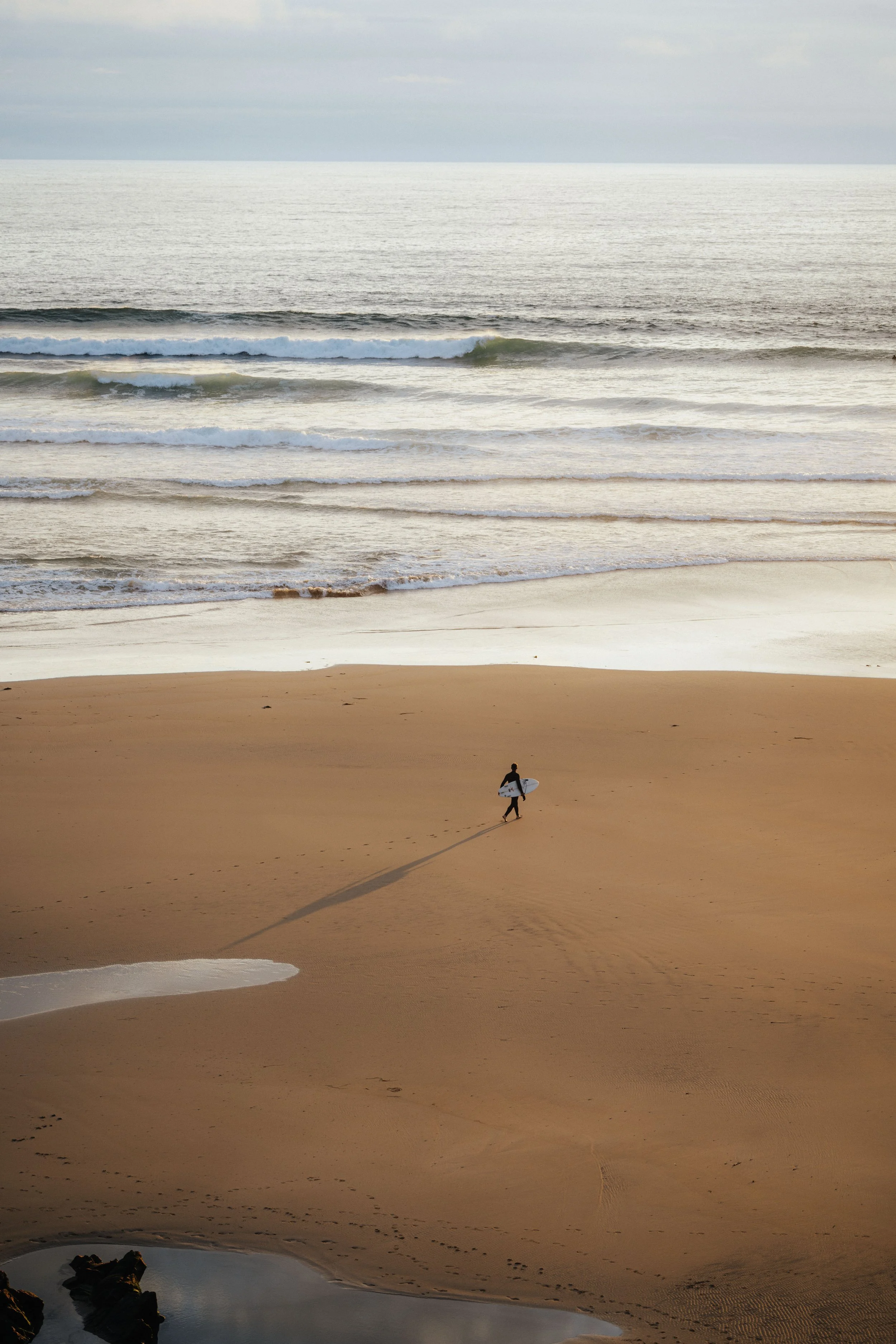 A solitary surfer walking on a sandy beach carrying a surfboard, with the ocean waves in the background.