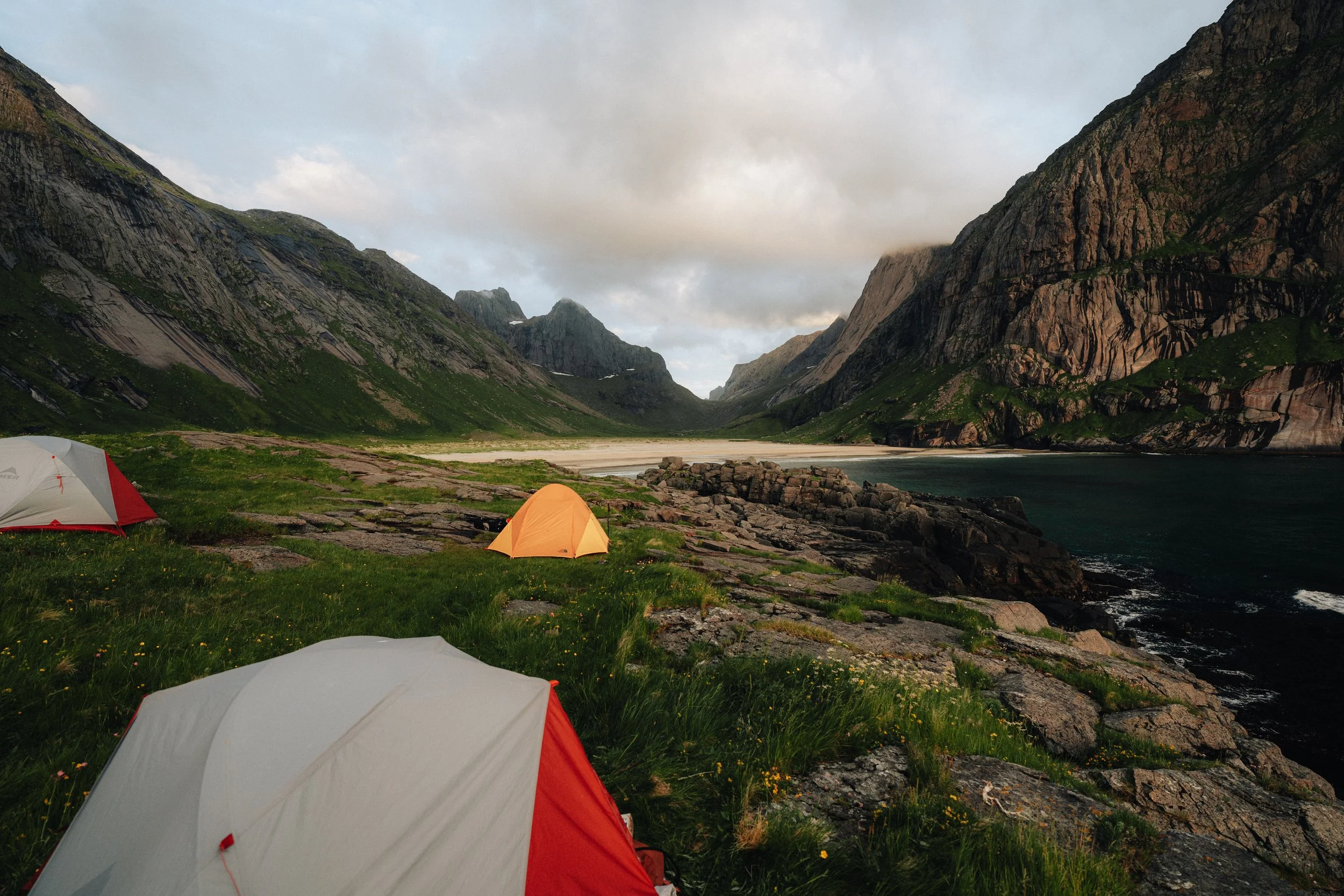 Three tents set up on grassy terrain near rocky shoreline with mountains in the background, under a cloudy sky.