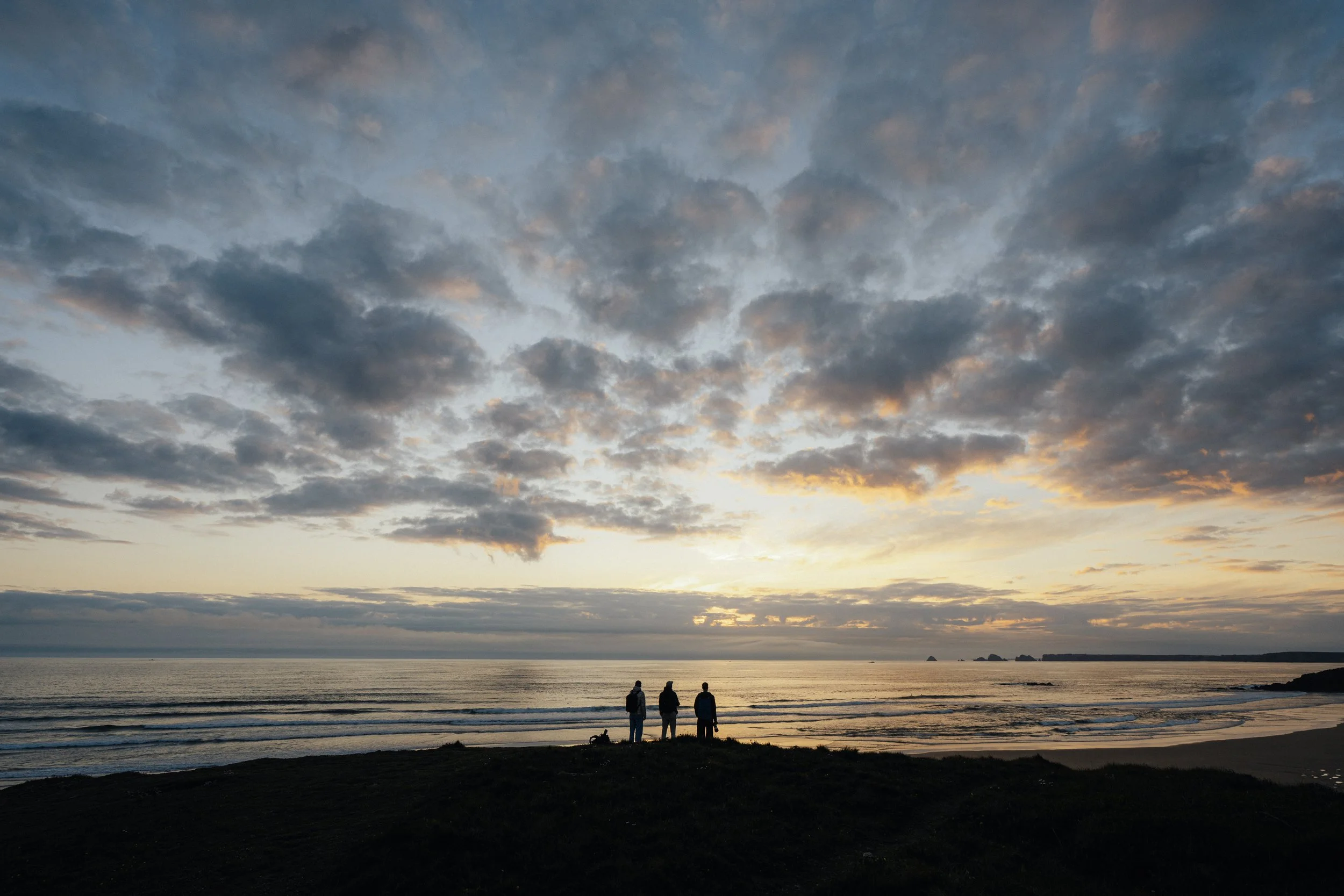 Three people standing on a beach during sunset, with clouds in the sky and the ocean in the background.