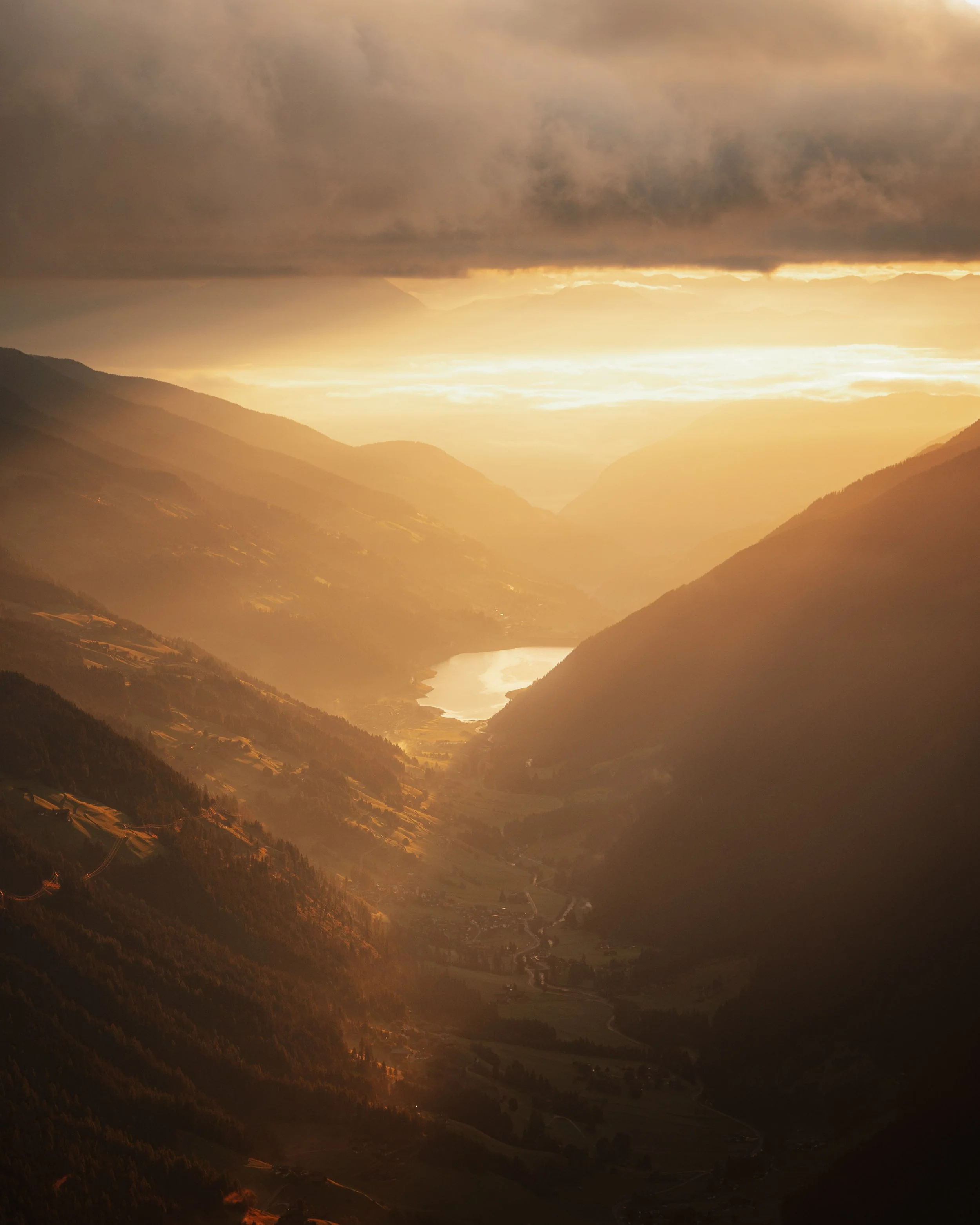 A scenic view of a mountain valley during sunset, with a river winding through the valley and distant mountains under a partly cloudy sky.