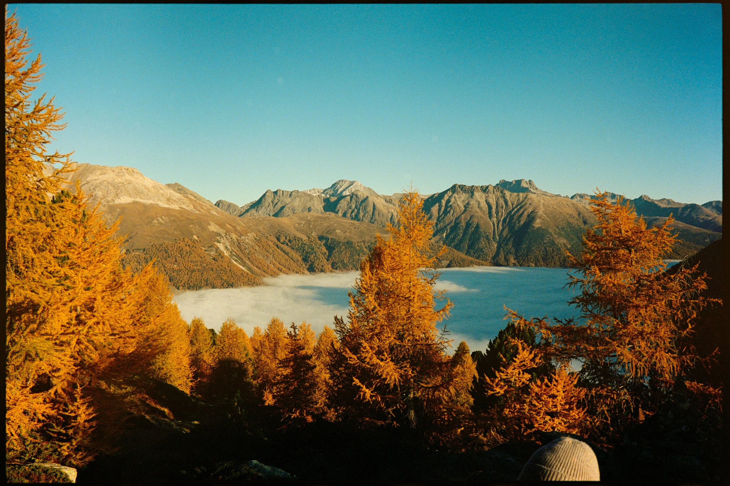 Scenic view of a mountain range with autumn-colored trees in the foreground and a lake surrounded by mountains and clouds in the background on a clear day.