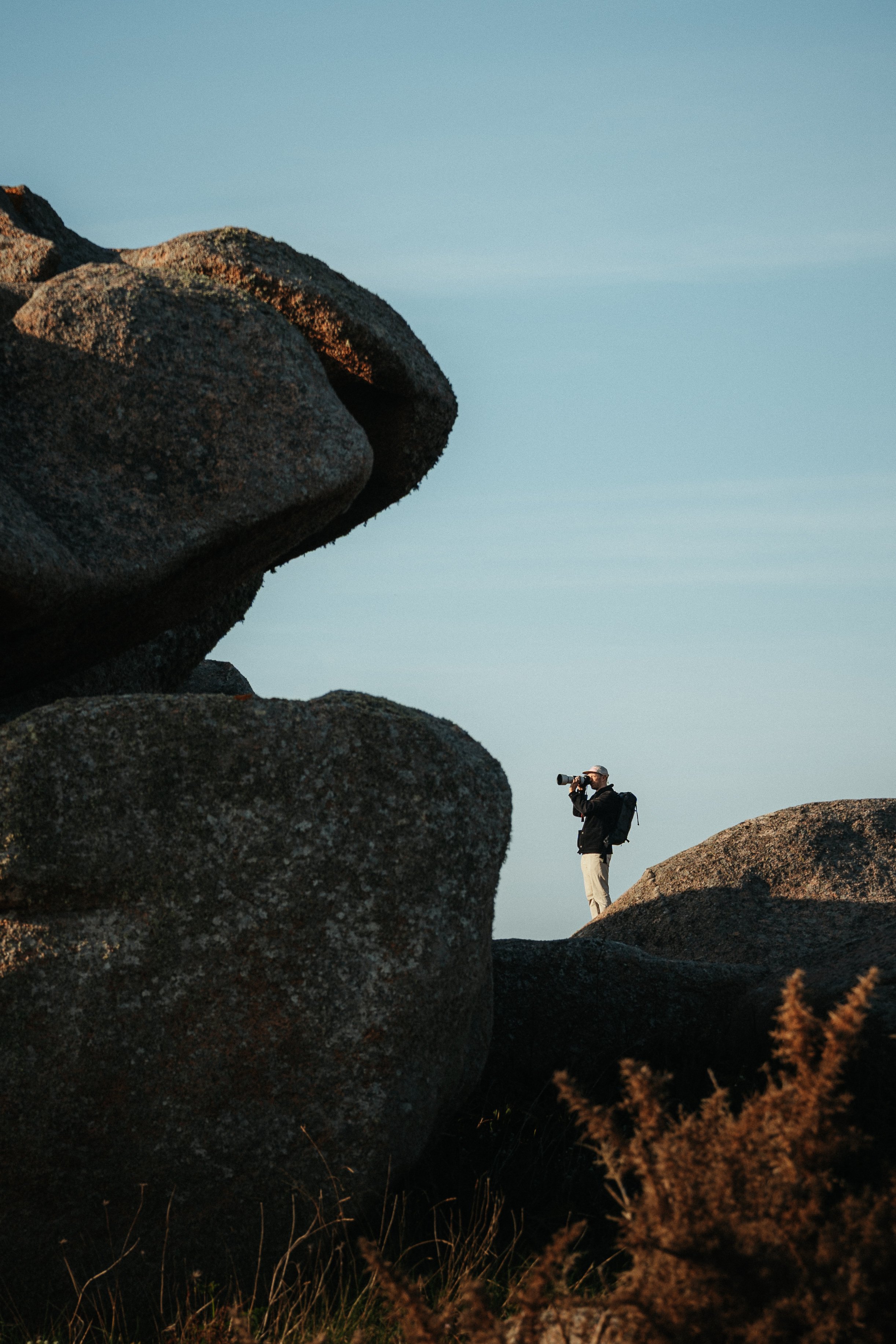 A person stands on a rocky terrain taking a photograph with a DSLR camera, surrounded by large rocks under a clear sky.