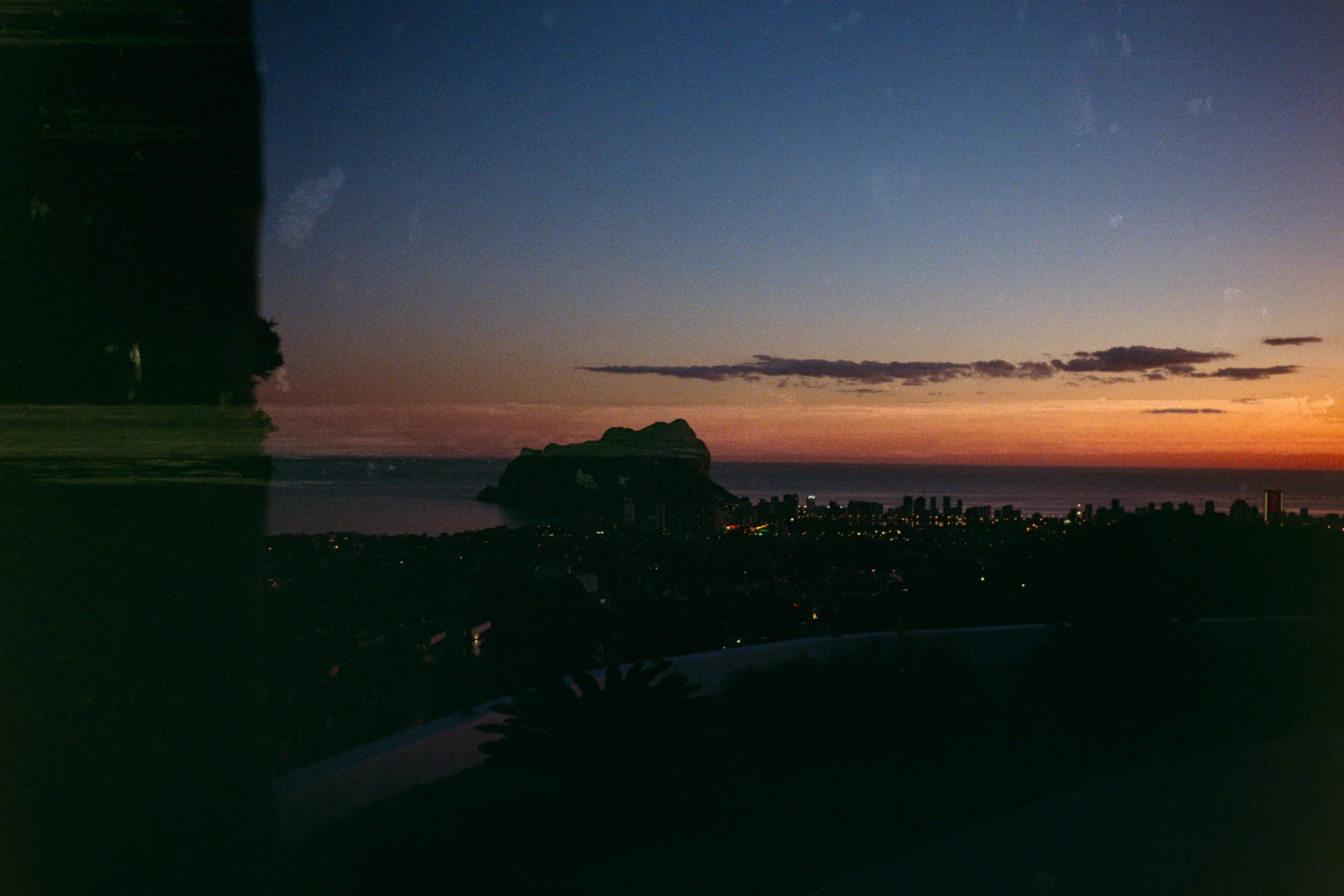Sunset over a city skyline with a large rock formation in the ocean, viewed through a window with reflections.