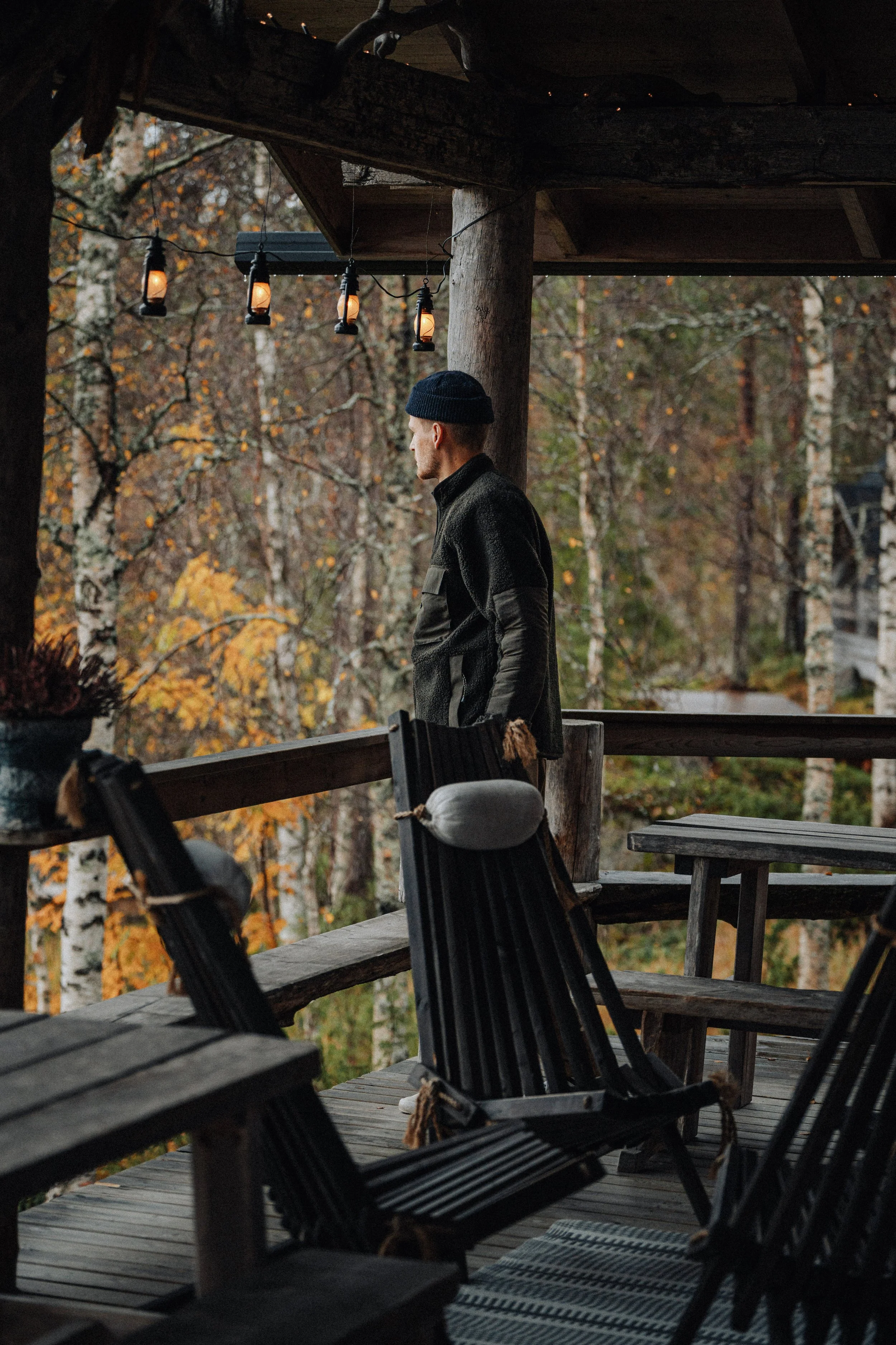 A man in a dark jacket and navy beanie stands on a wooden deck surrounded by autumn trees, looking out into the forest. String lights hang above the deck, which also features wooden outdoor furniture, including chairs with pillows. The scene appears 