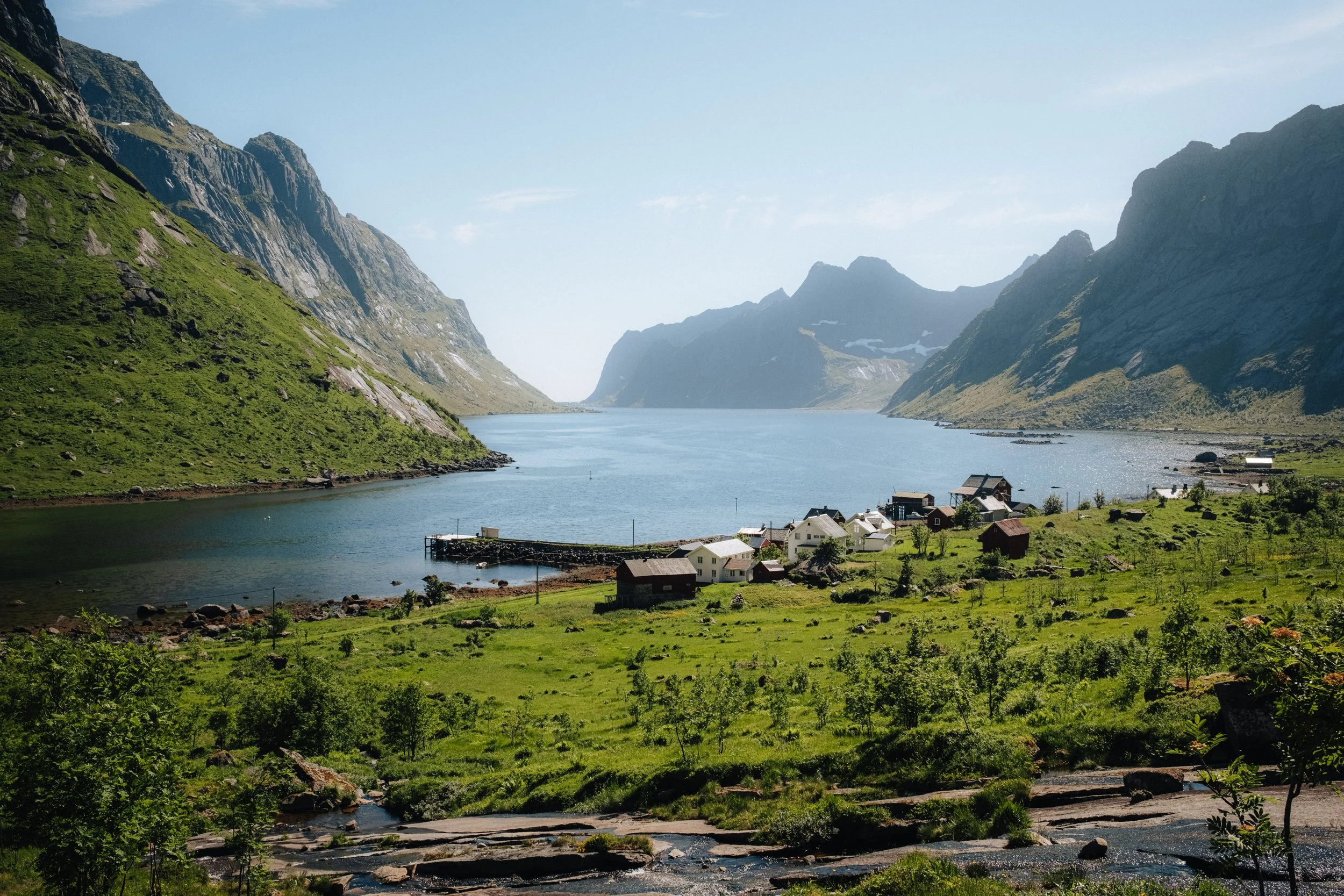 A scenic view of a small village by a lake, surrounded by green mountains under a clear sky.