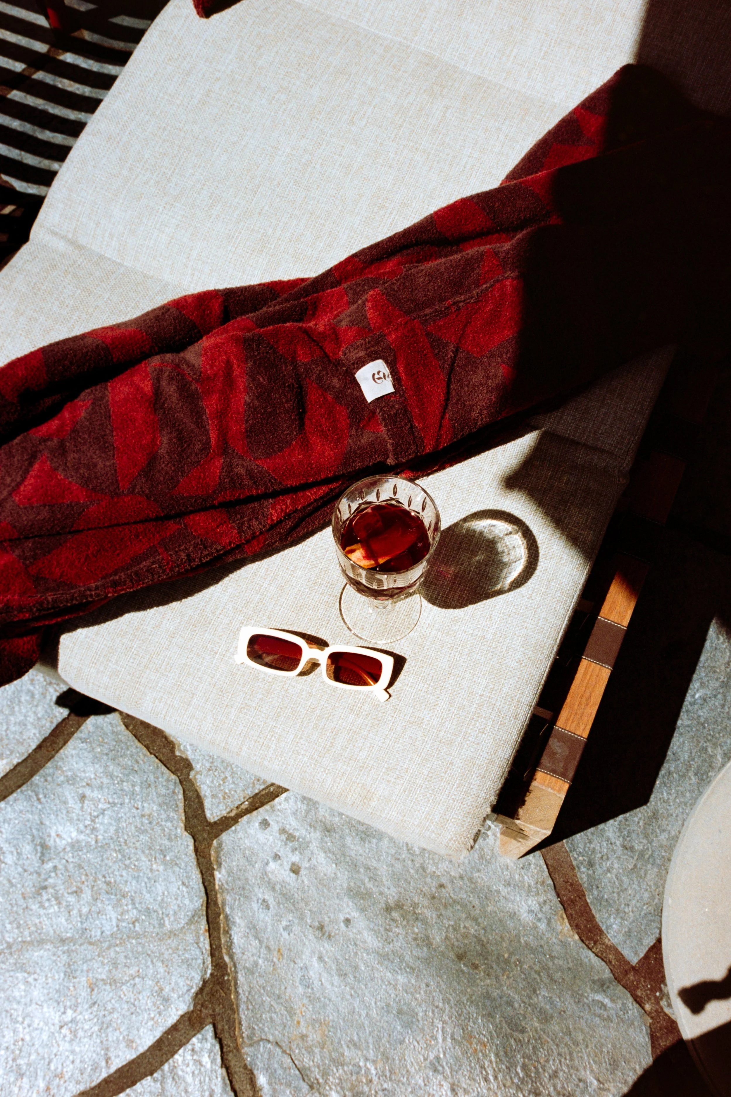 A beige outdoor chair with a red plaid blanket, sunglasses with white frames, and a glass of dark beverage. The chair is on a stone patio, and sunlight casts shadows of the drink and sunglasses on the chair.