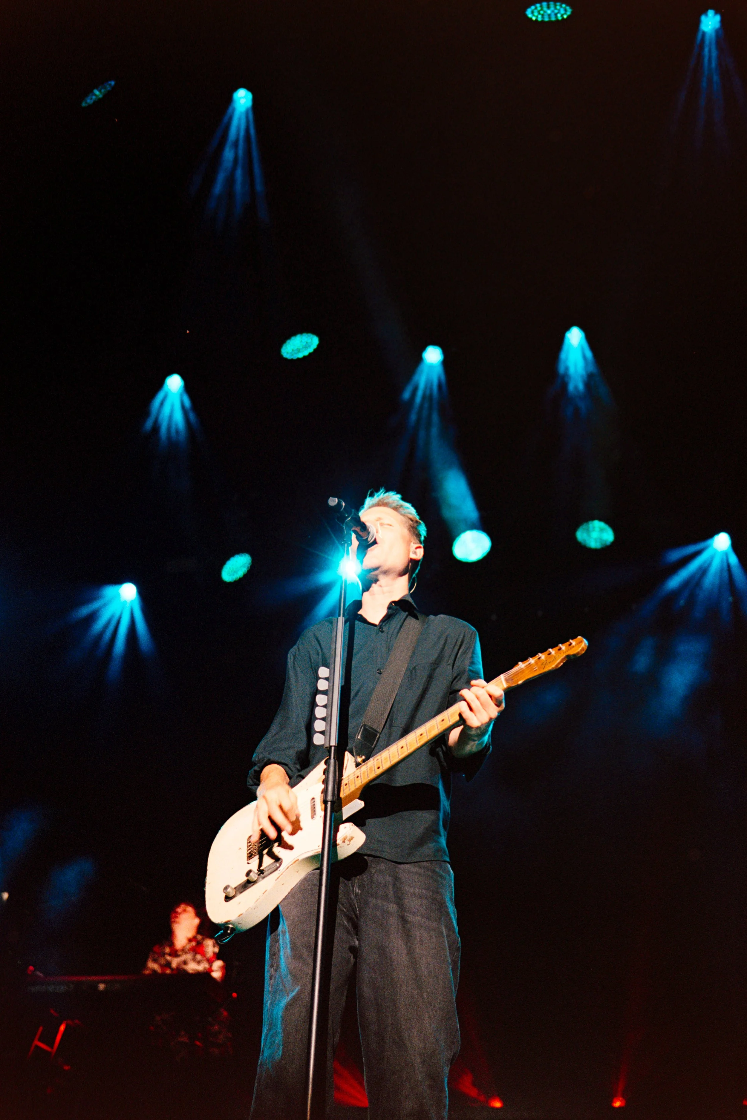 A musician performing on stage with a white electric guitar, singing into a microphone, with blue stage lights overhead and a keyboard player in the background.