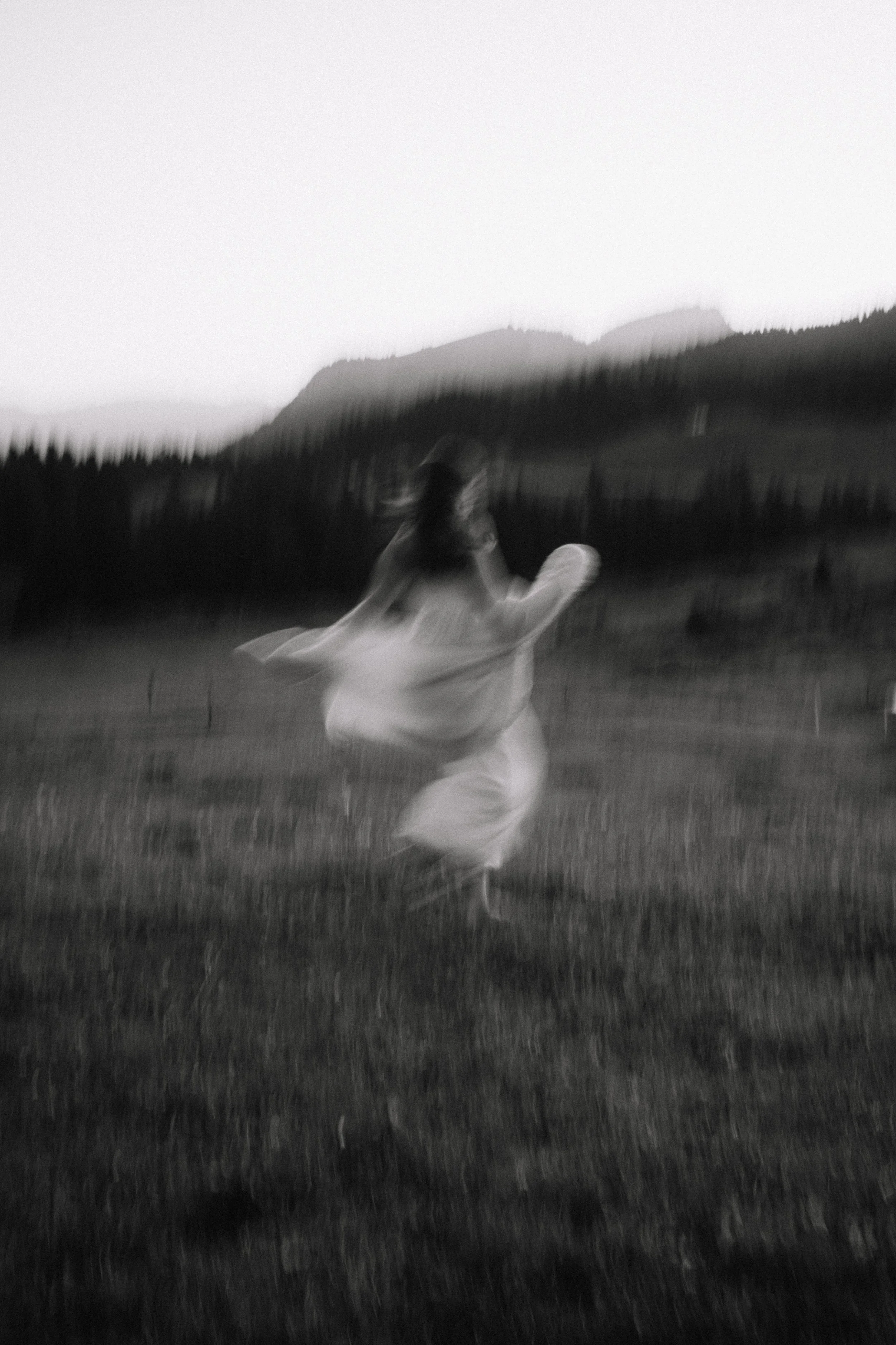 A black and white photo of a woman twirling in a field with mountains in the background, captured with motion blur.