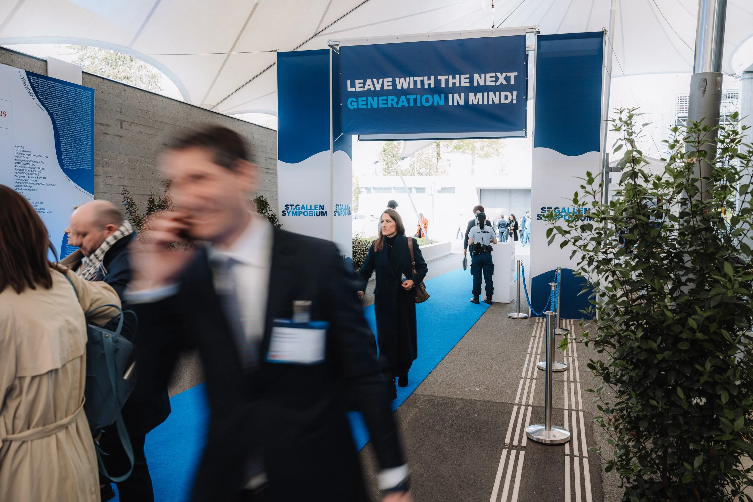 Entrance to the St. Gallen Symposium with a blue sign that reads "Leave with the next generation in mind!" and attendees walking through the corridor.