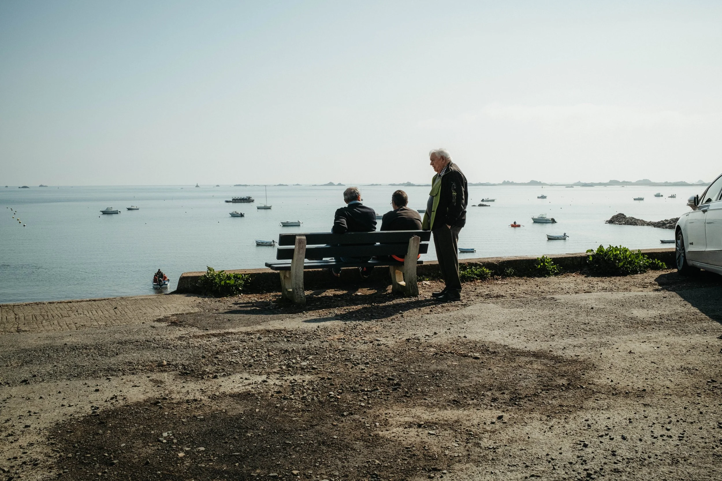 Three men near a waterfront, with two sitting on a bench and one standing, while looking at boats in the water.