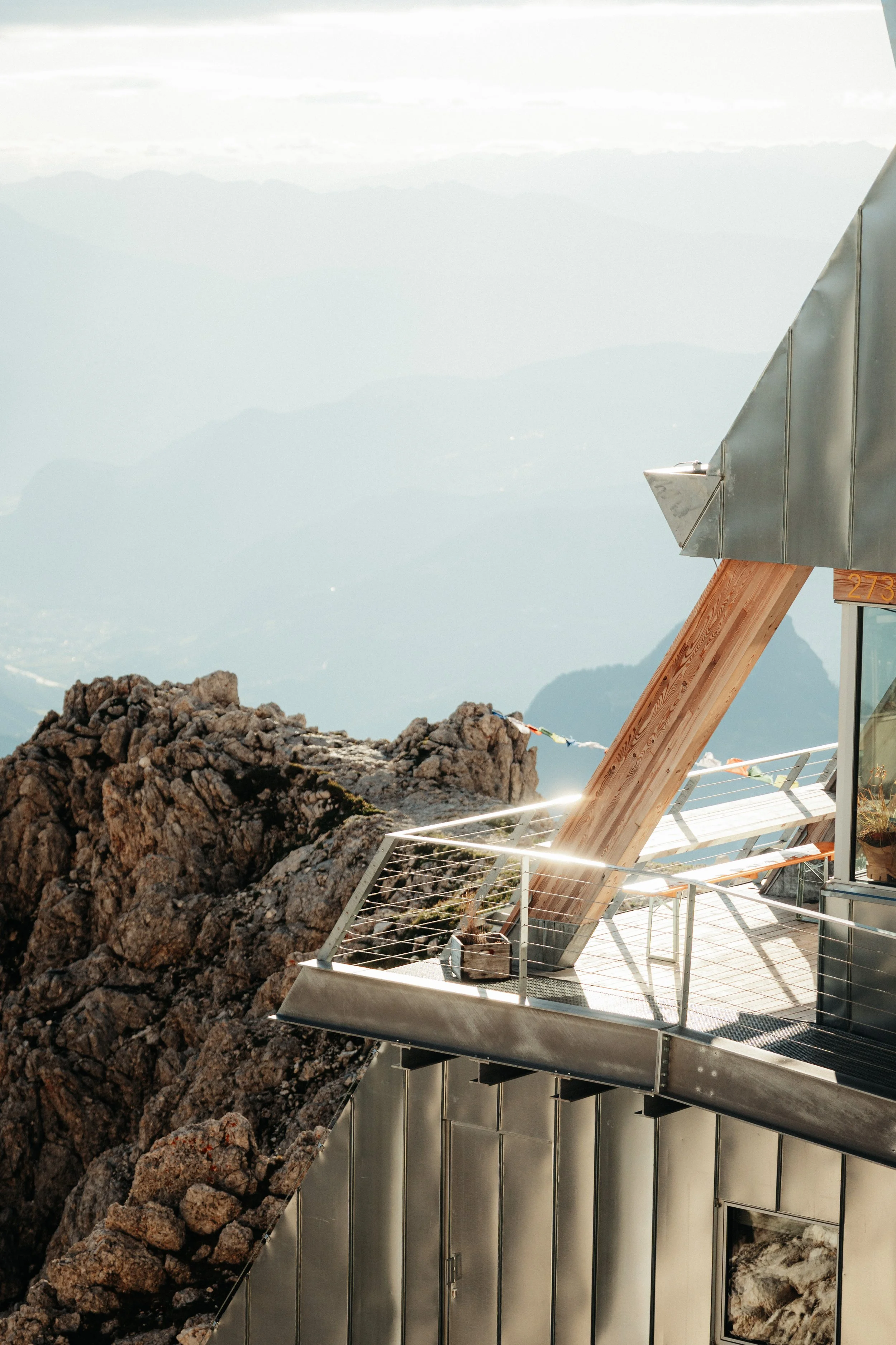 A mountain observation deck with a metal and wooden structure, overlooking rocky terrain and distant mountain ranges.
