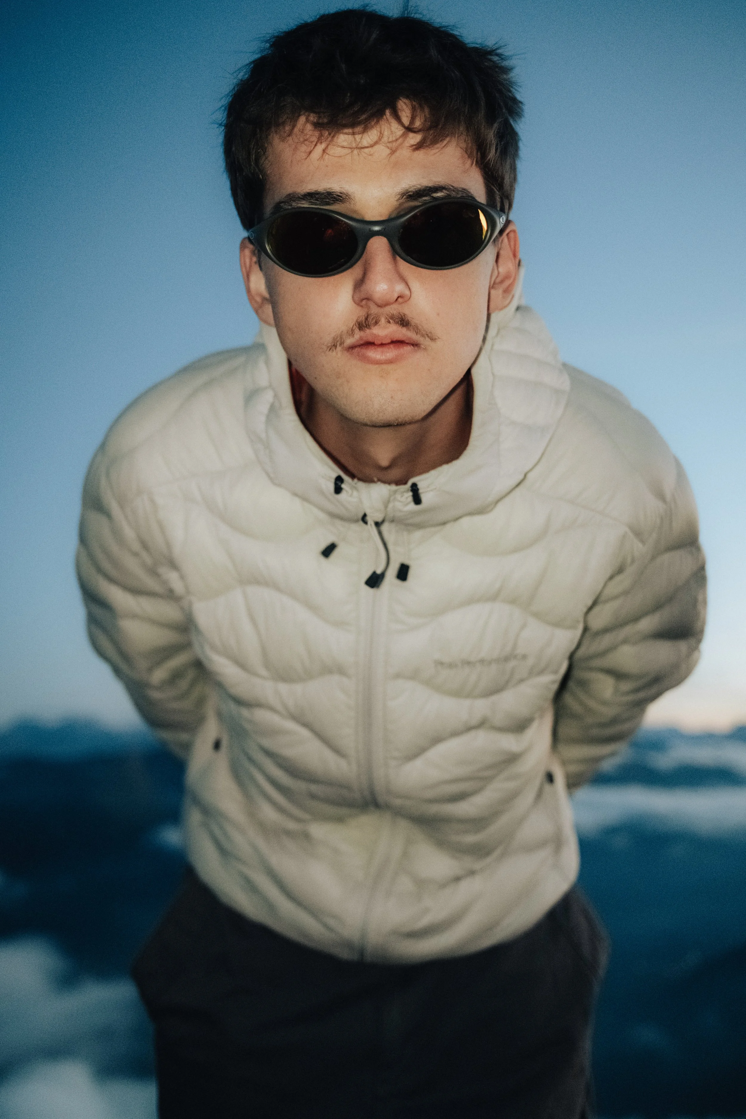 A young man with dark hair, wearing black sunglasses and a white puffy jacket, leaning forward with a mountainous landscape in the background.