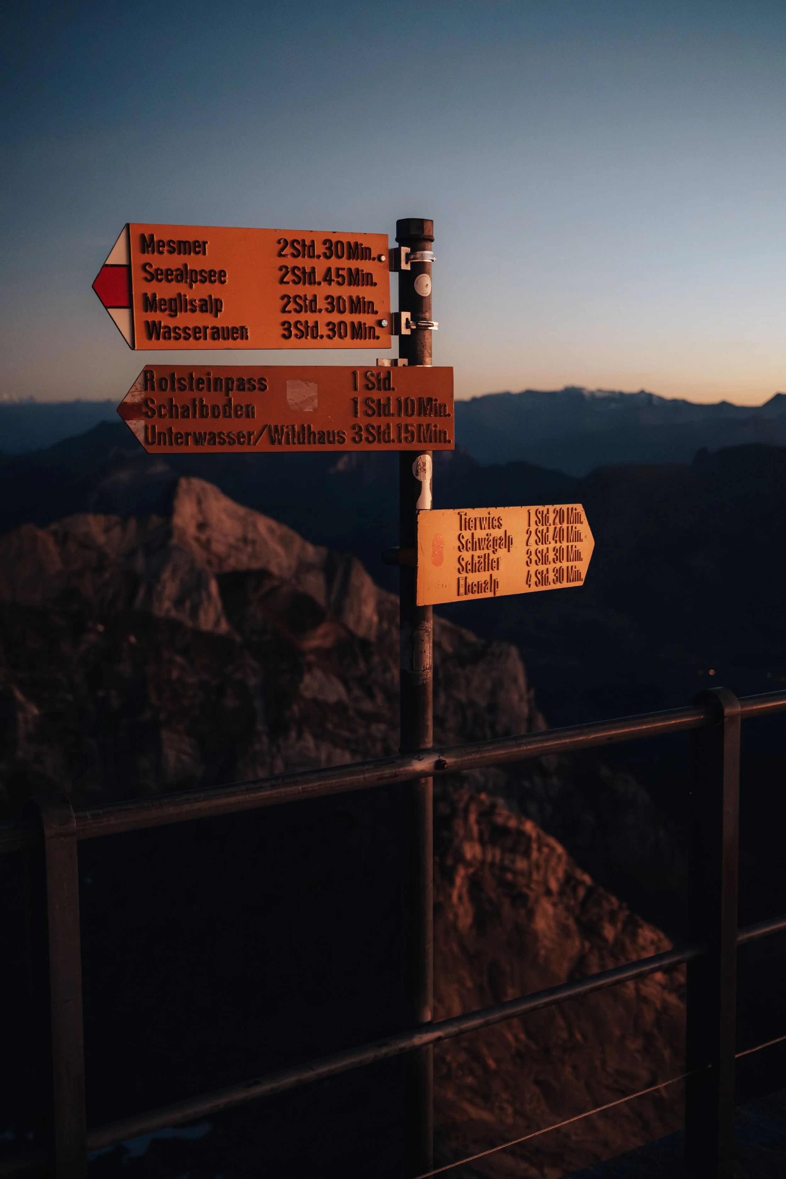 Mountain trail sign with multiple rows of orange and brown directional signs at dusk, pointing towards different mountain peaks and routes, with a mountain range and sunset in the background.