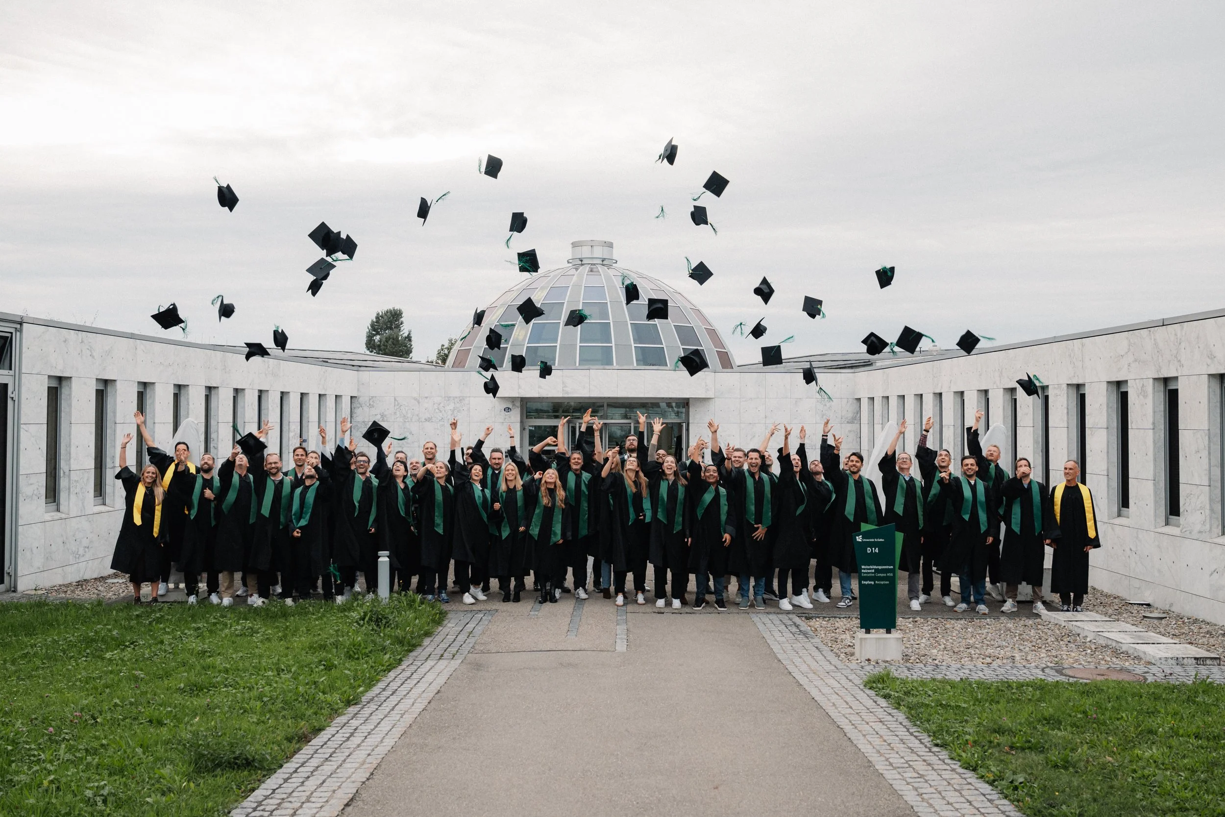 A group of graduates in caps and gowns celebrating their graduation outdoors with caps tossed in the air in front of a modern building with a glass dome on overcast day.