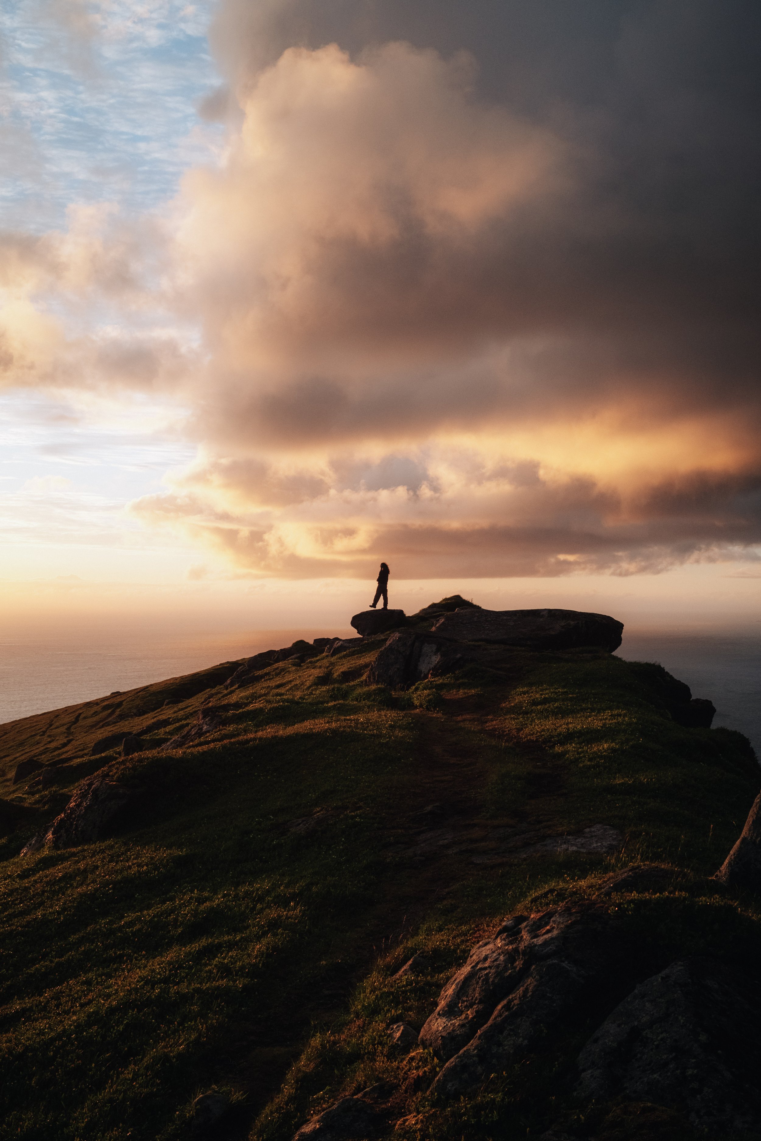 A silhouette of a person standing on a rock formation atop a grassy hill, with dramatic clouds and a sunset sky in the background