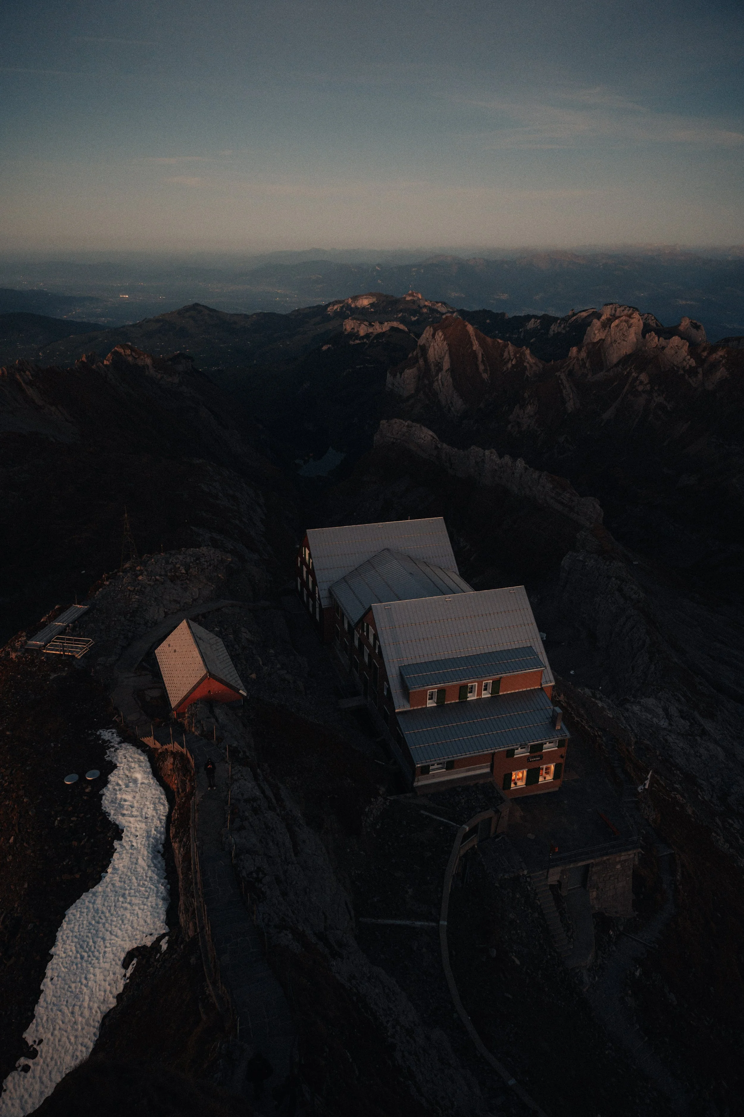 Aerial view of mountain landscape at dusk with a large building and smaller structures on a rocky ridge.
