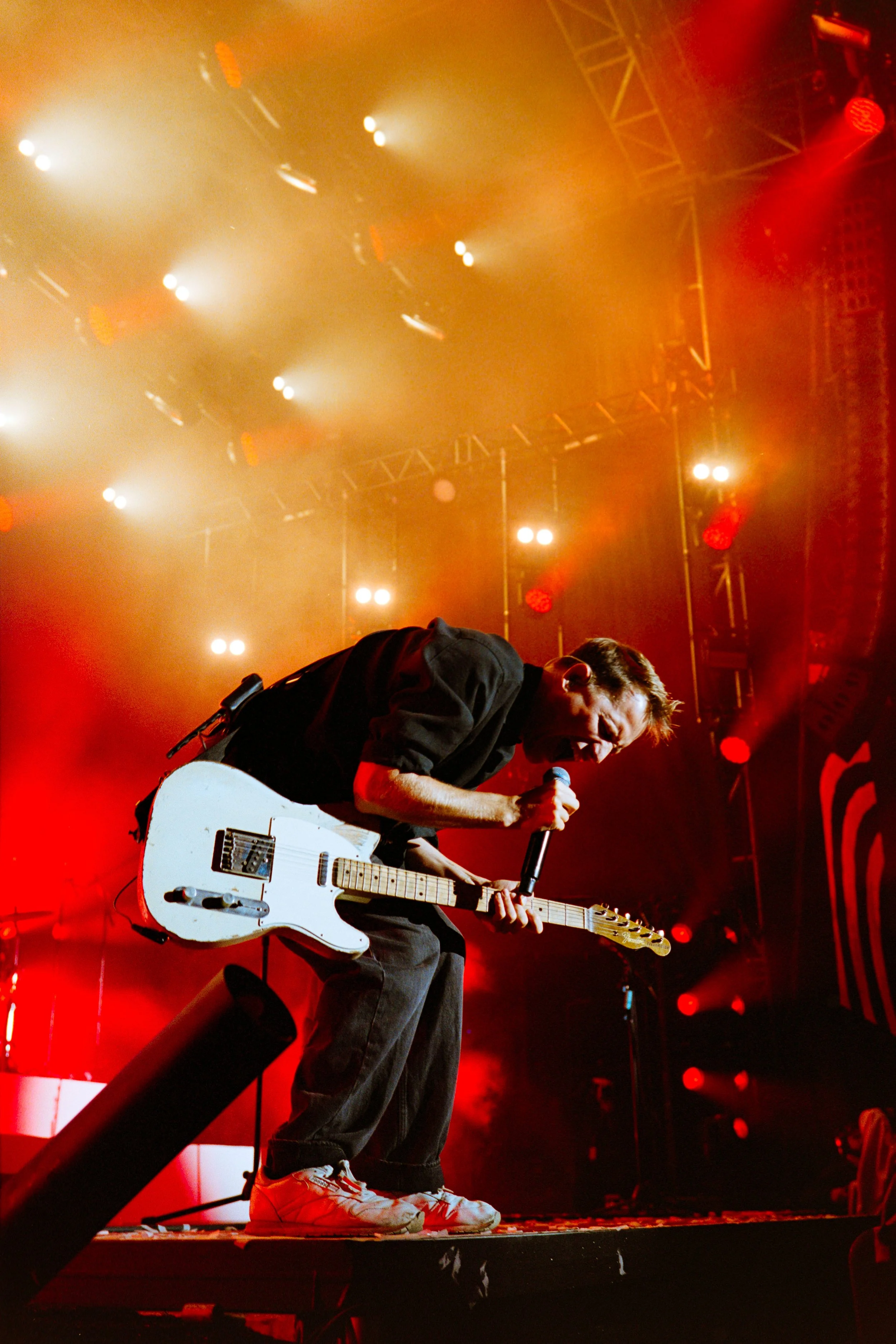 Musician playing an electric guitar and singing into a microphone on stage, with bright lighting and red stage lights.