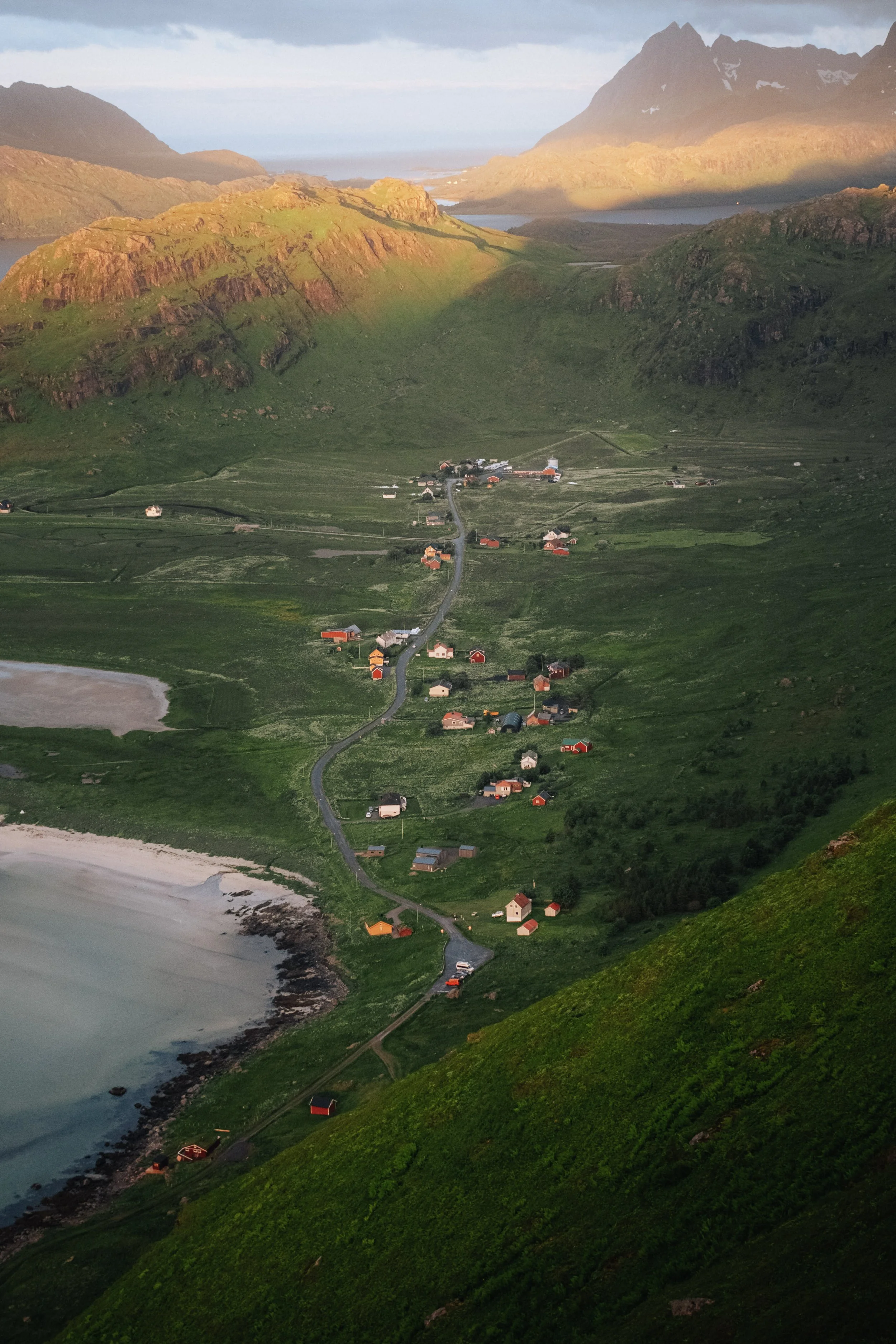 A mountainous landscape with a small village, green hills, a beach, and a calm sea, lit by warm sunlight.