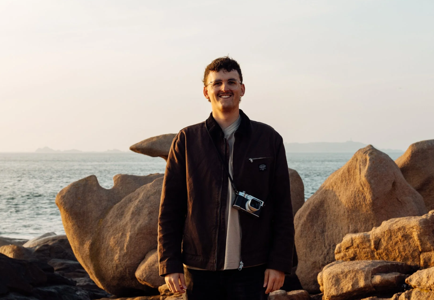 A smiling man in a black jacket with a camera hanging from his neck stands on a rocky beach with the ocean in the background during sunset.