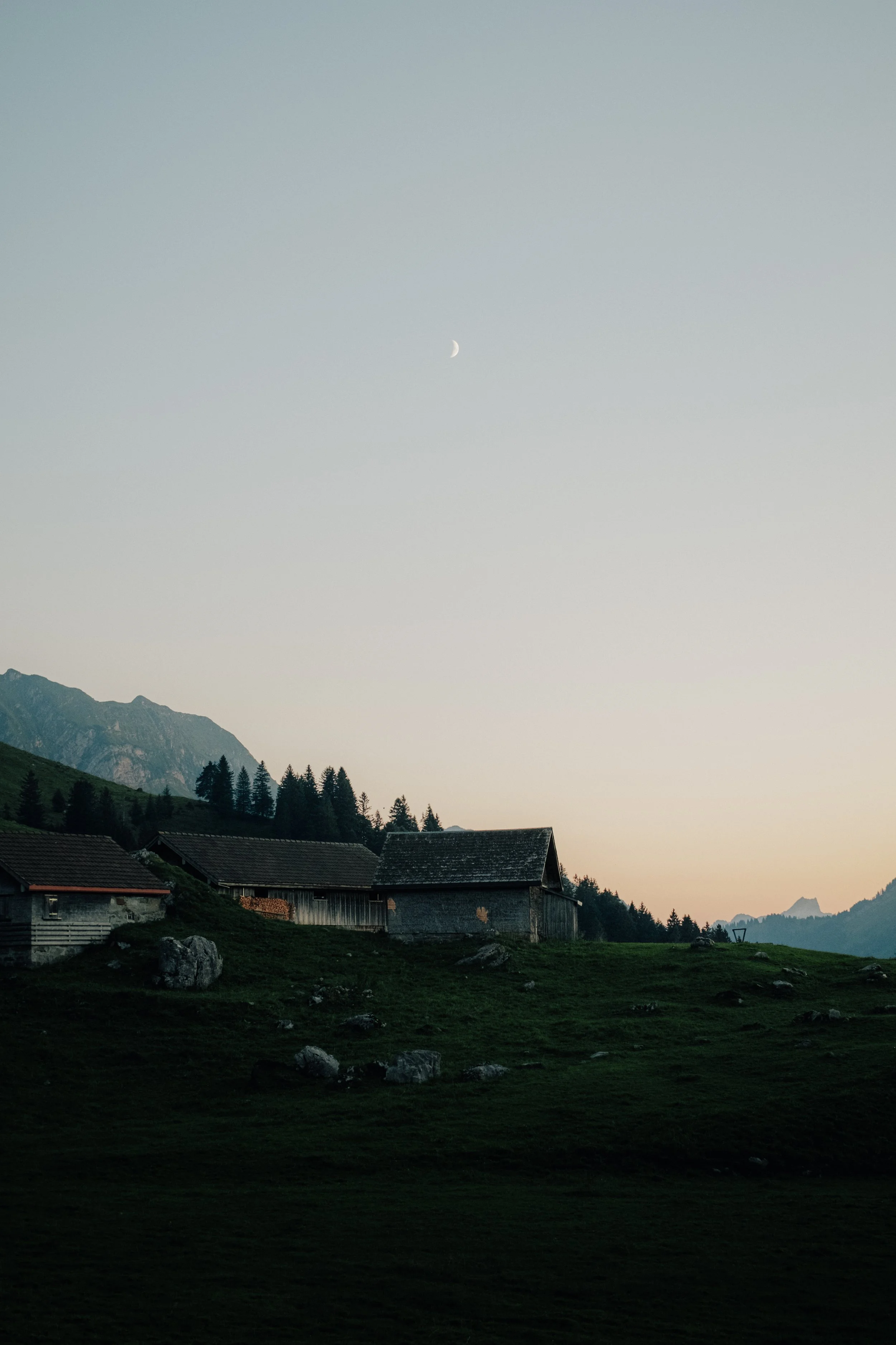 A scenic landscape at dusk with remote wooden cabins on a grassy hill, mountains in the background, a clear sky with a crescent moon, and trees along the hills.