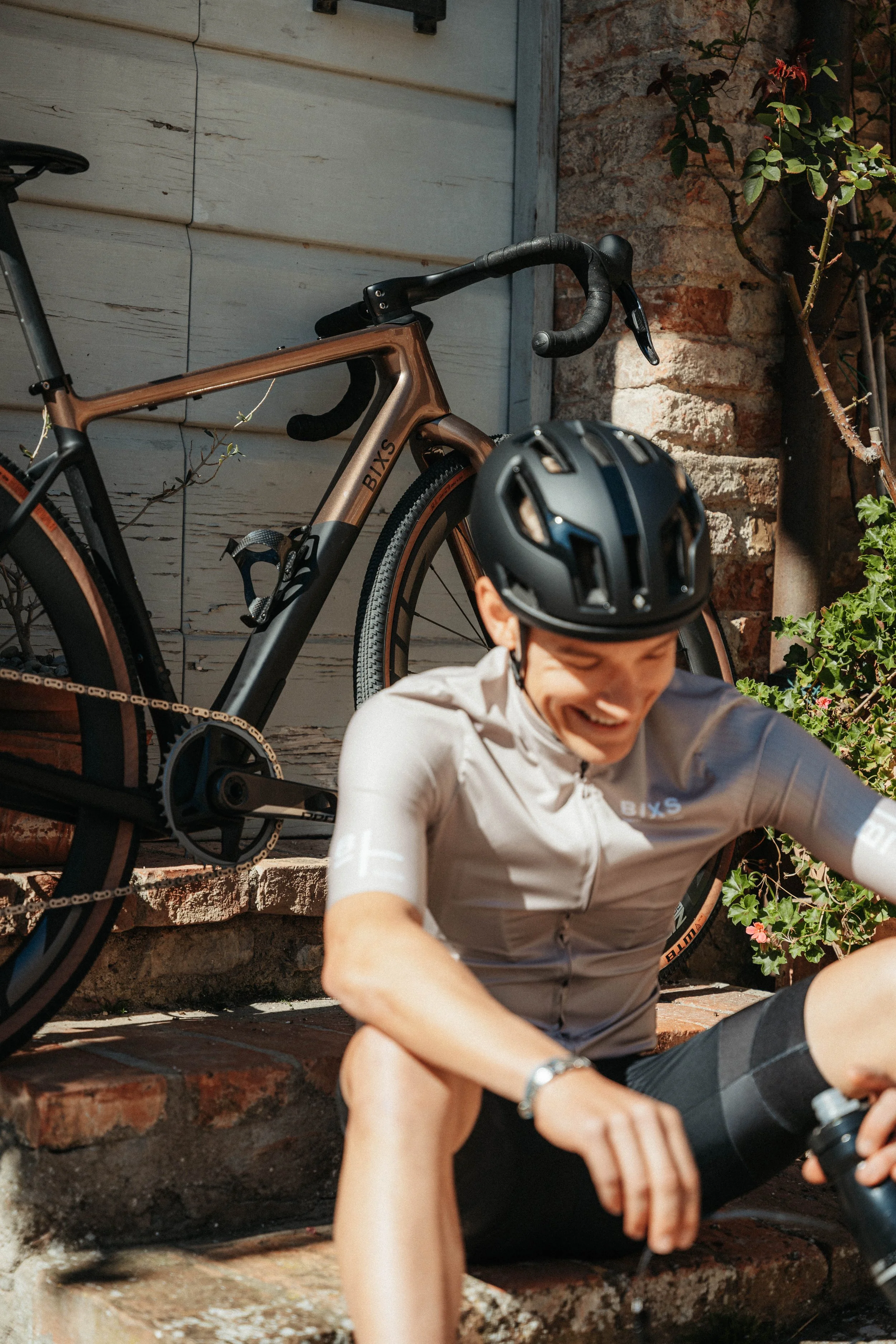 A man wearing a helmet and cycling outfit sits on brick steps, smiling and adjusting his shoe, with a bicycle parked behind him against a wall.