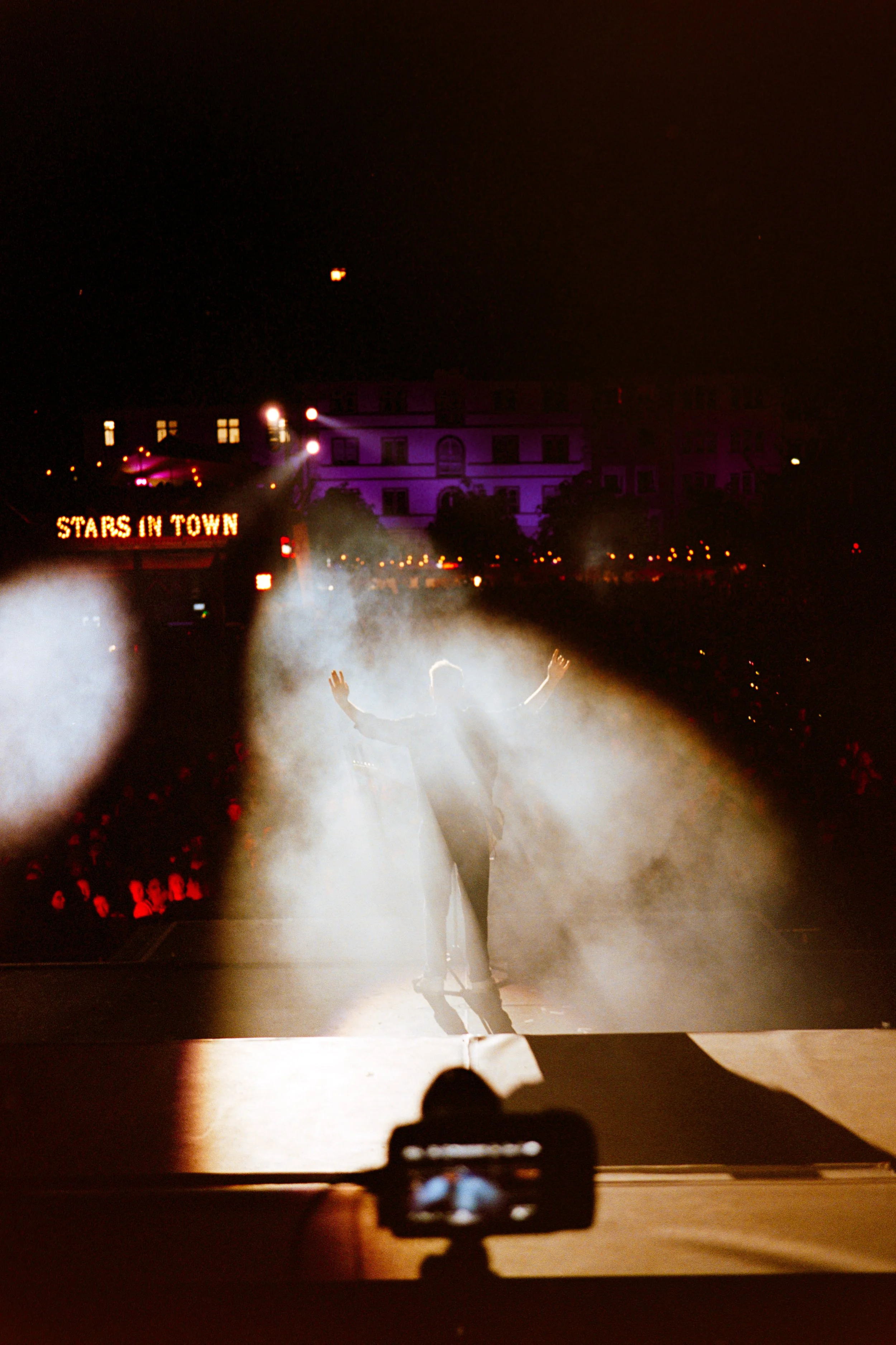 Performer on stage with arms raised, illuminated by spotlight, in front of an audience at night, with a sign reading 'Stars in Town' in the background.