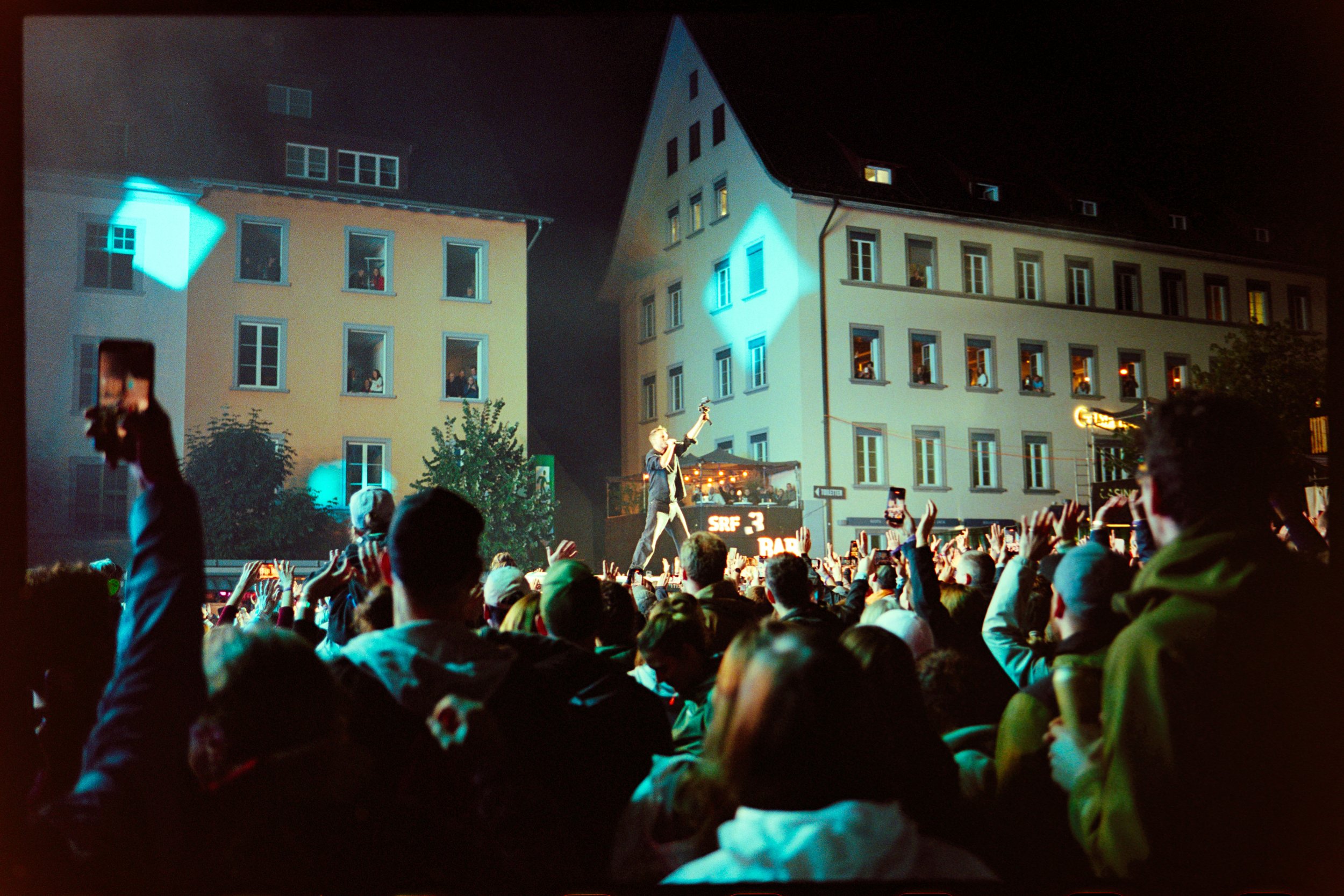A concert in an outdoor urban setting at night with a crowd of people watching a performer on stage. The stage is in front of buildings, and the performer is holding a microphone and engaging with the audience.