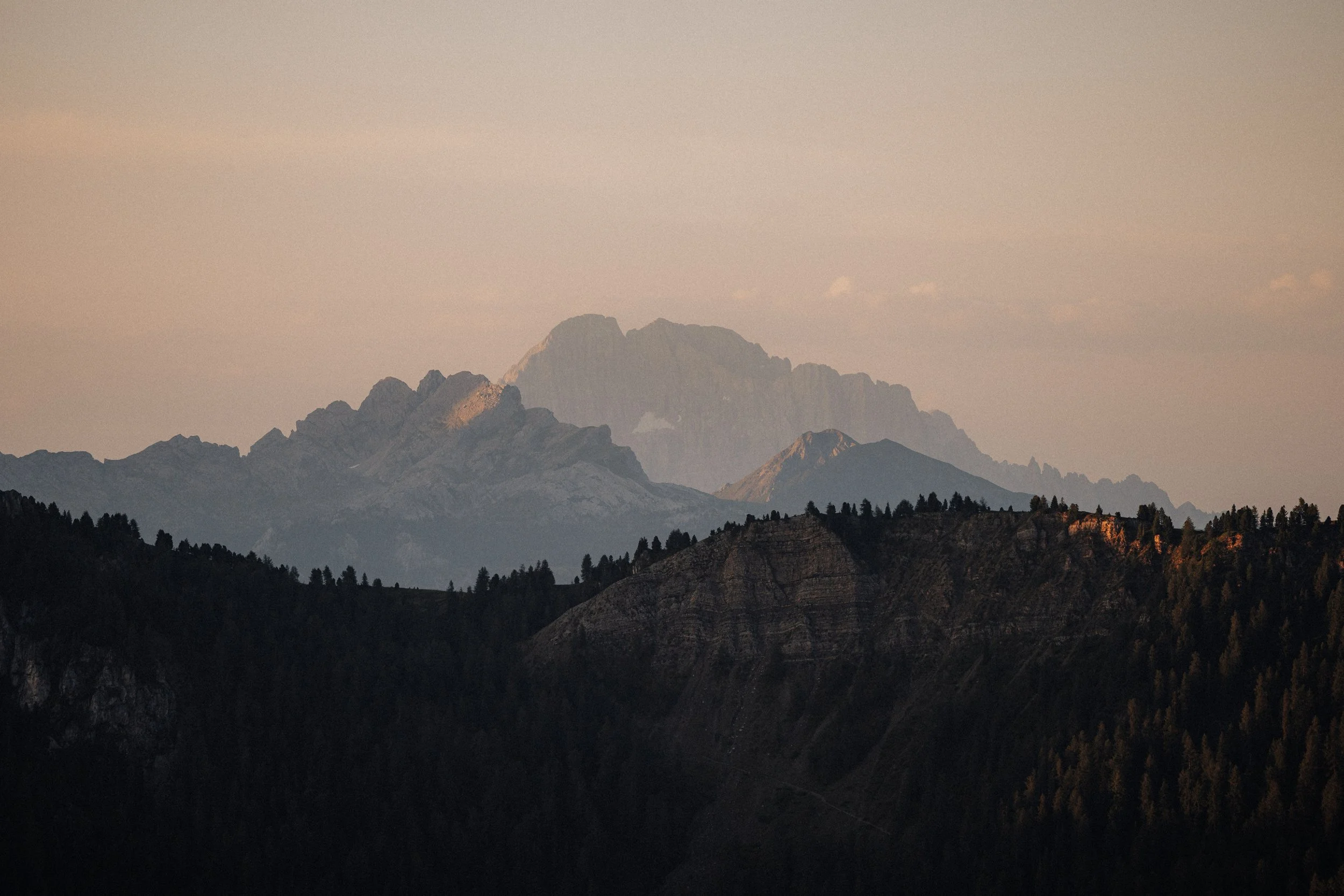 Mountain range with rugged peaks in the distance, layered with darker forested hills in the foreground under a pale sky.