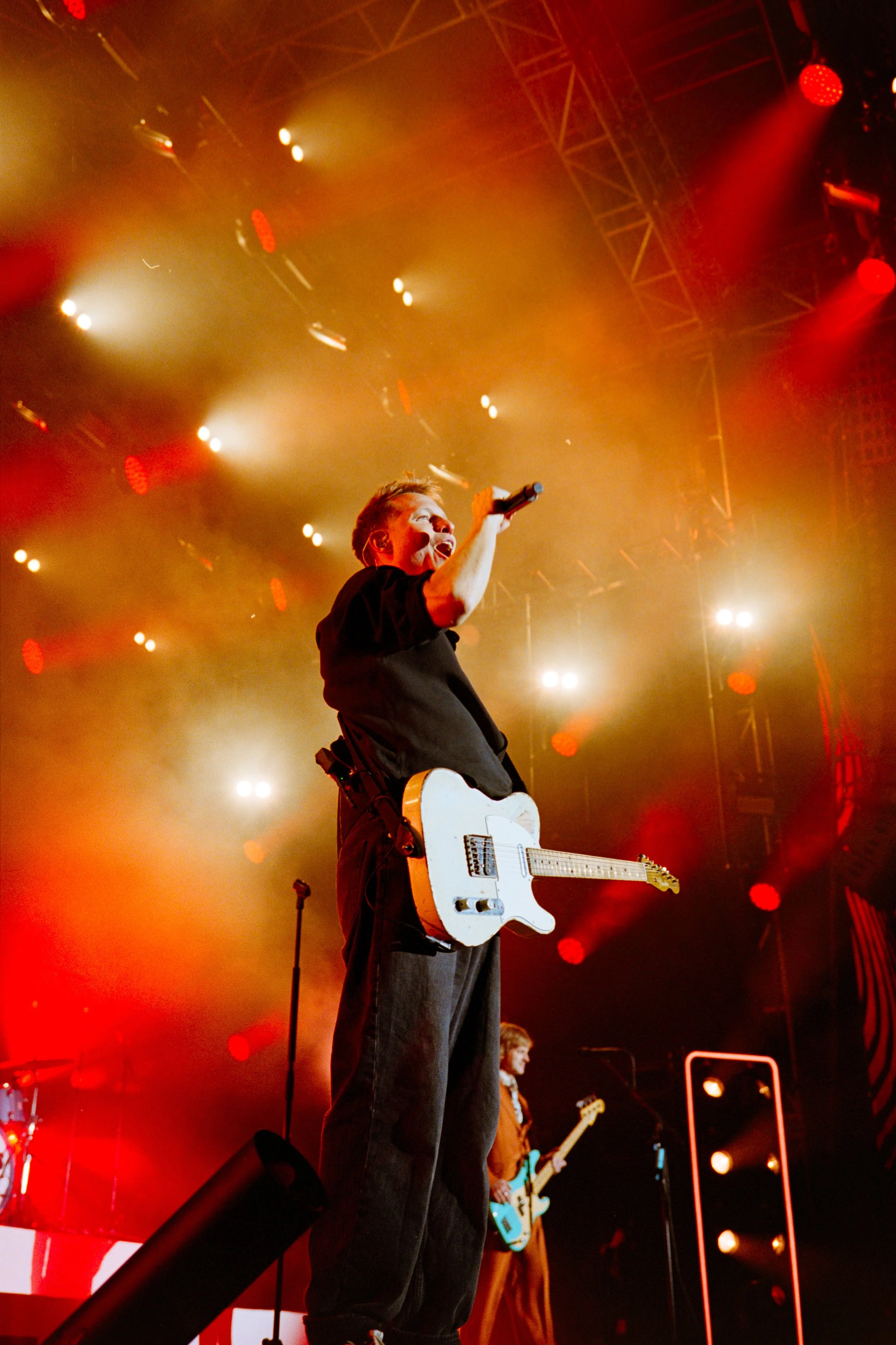Musician on stage singing into a microphone with a guitar around his neck, illuminated by colorful stage lights, with another band member playing guitar in the background.