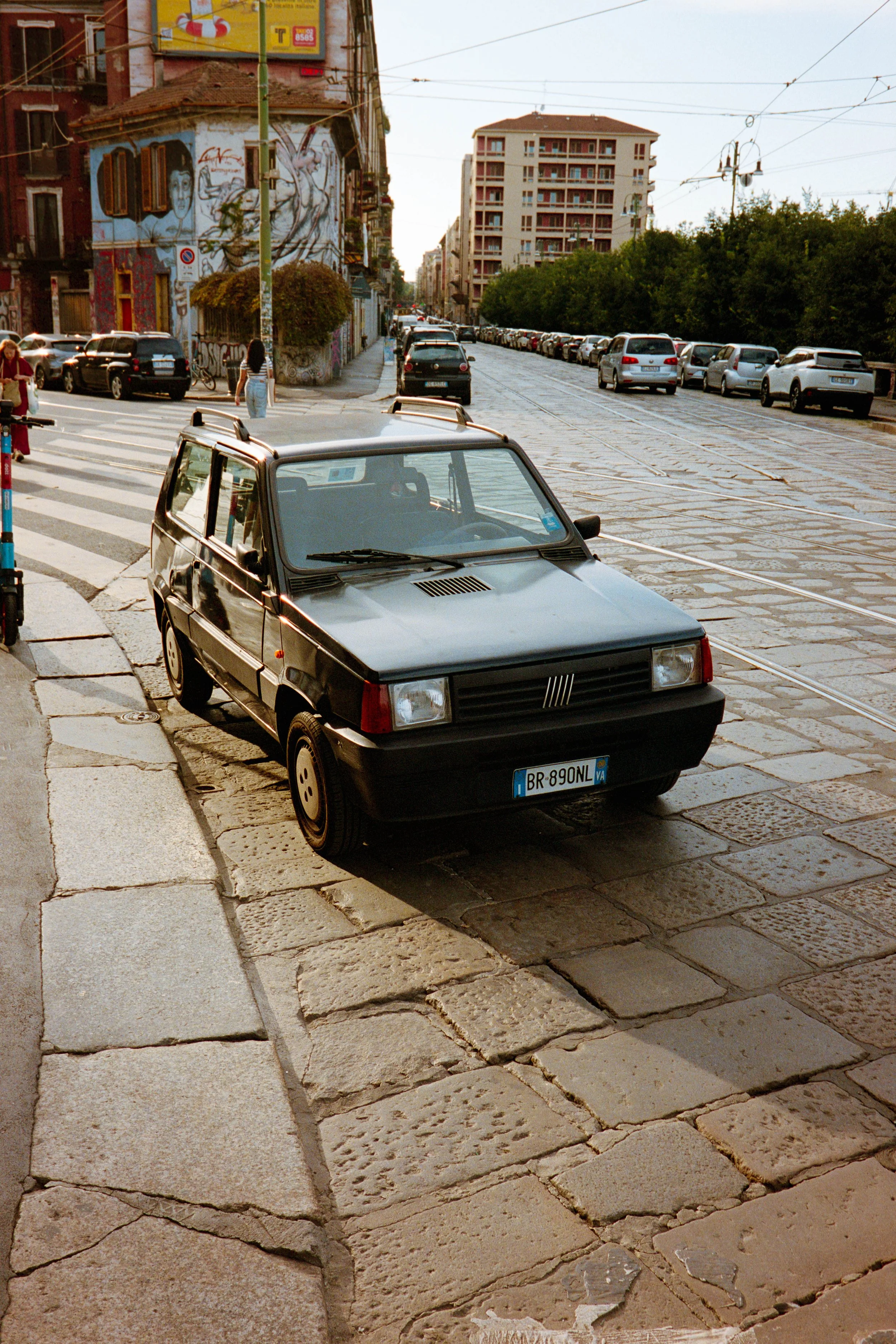 A black vintage Fiat parked on a cobblestone street in an urban area with buildings, trees, and parked cars visible in the background.
