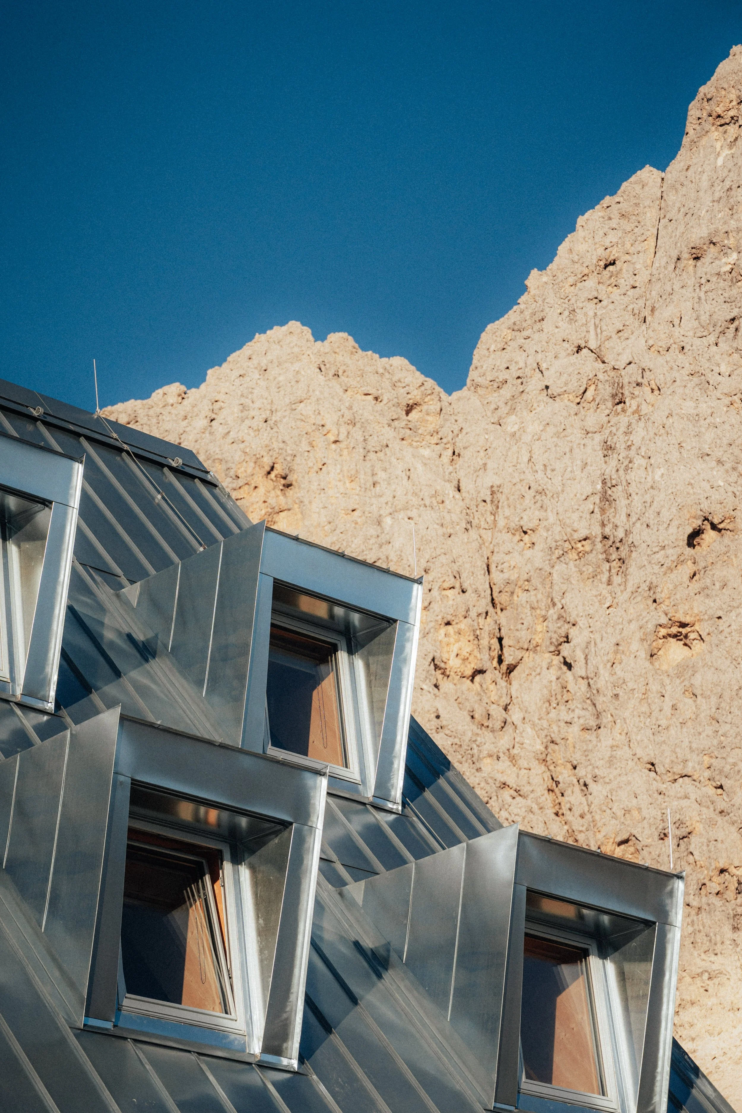 Close-up of modern building with metal roof and skylight windows, with a beige rocky mountain and clear blue sky in the background.