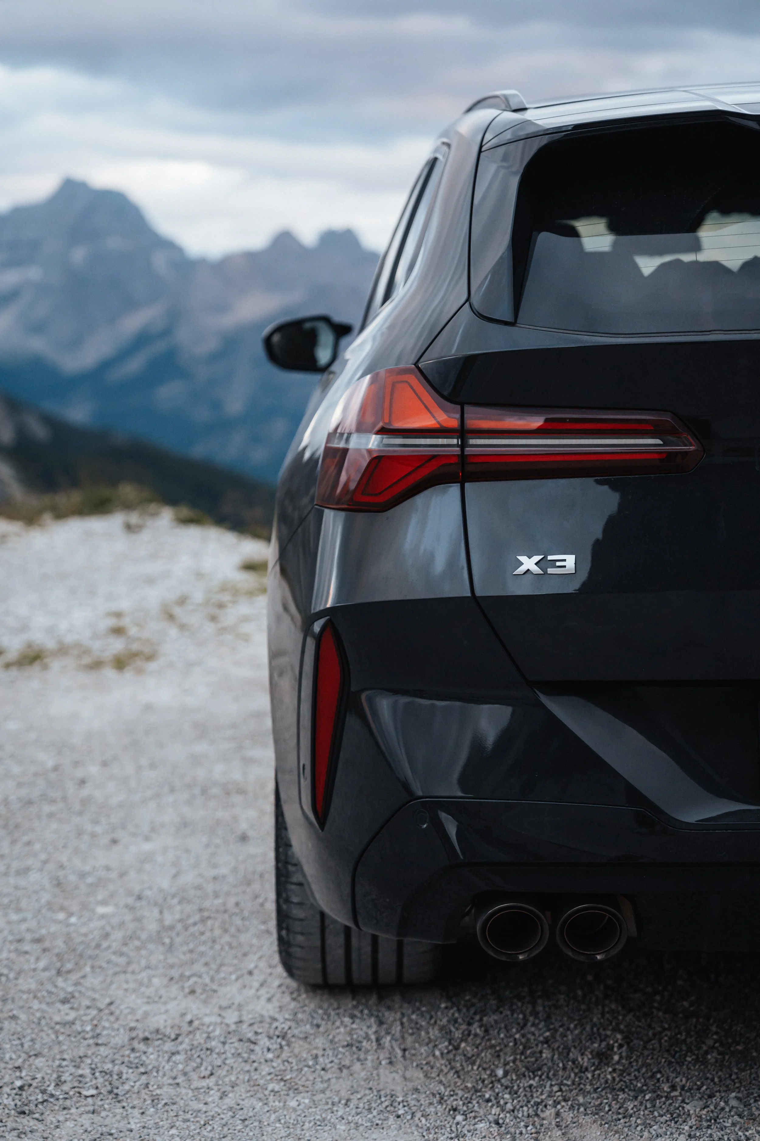 Close-up of the rear of a black Audi X3 SUV parked on a gravel surface with mountains in the background.