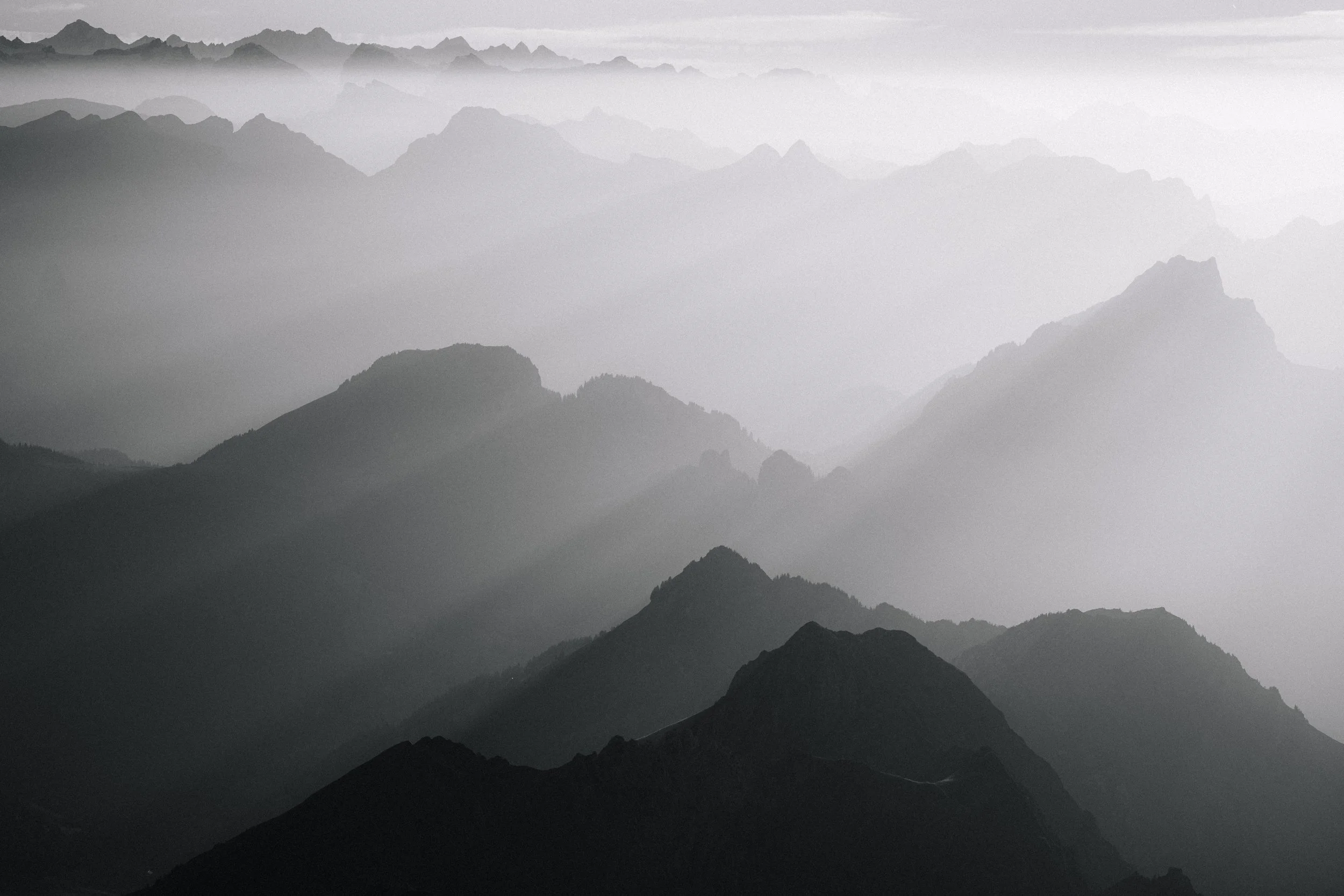 A black and white photo of layered mountain ranges shrouded in mist or fog, creating a sense of depth and mystery.