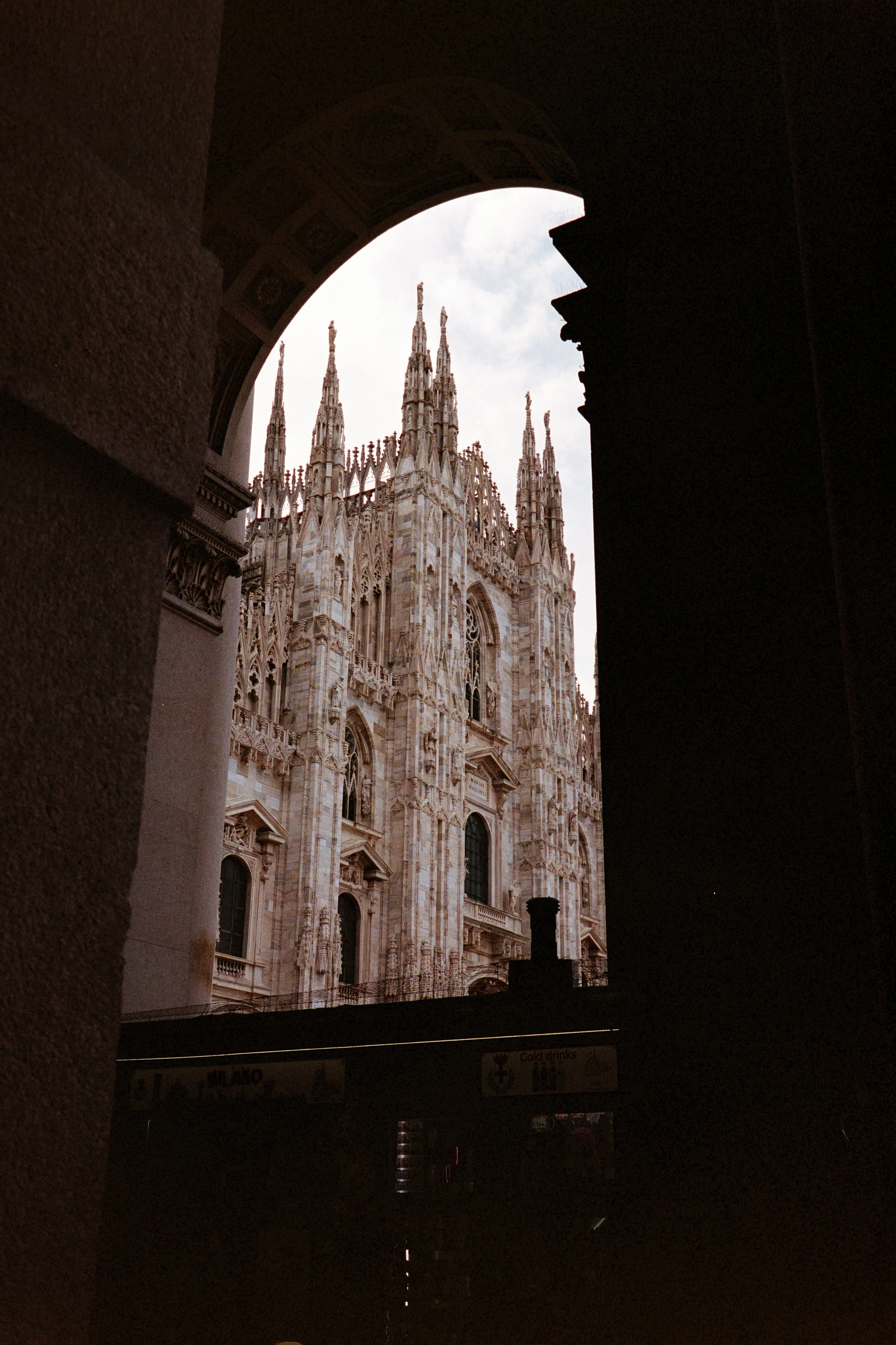 View of Milan Cathedral through an archway, showing its intricate Gothic architecture with tall spires and detailed stonework.