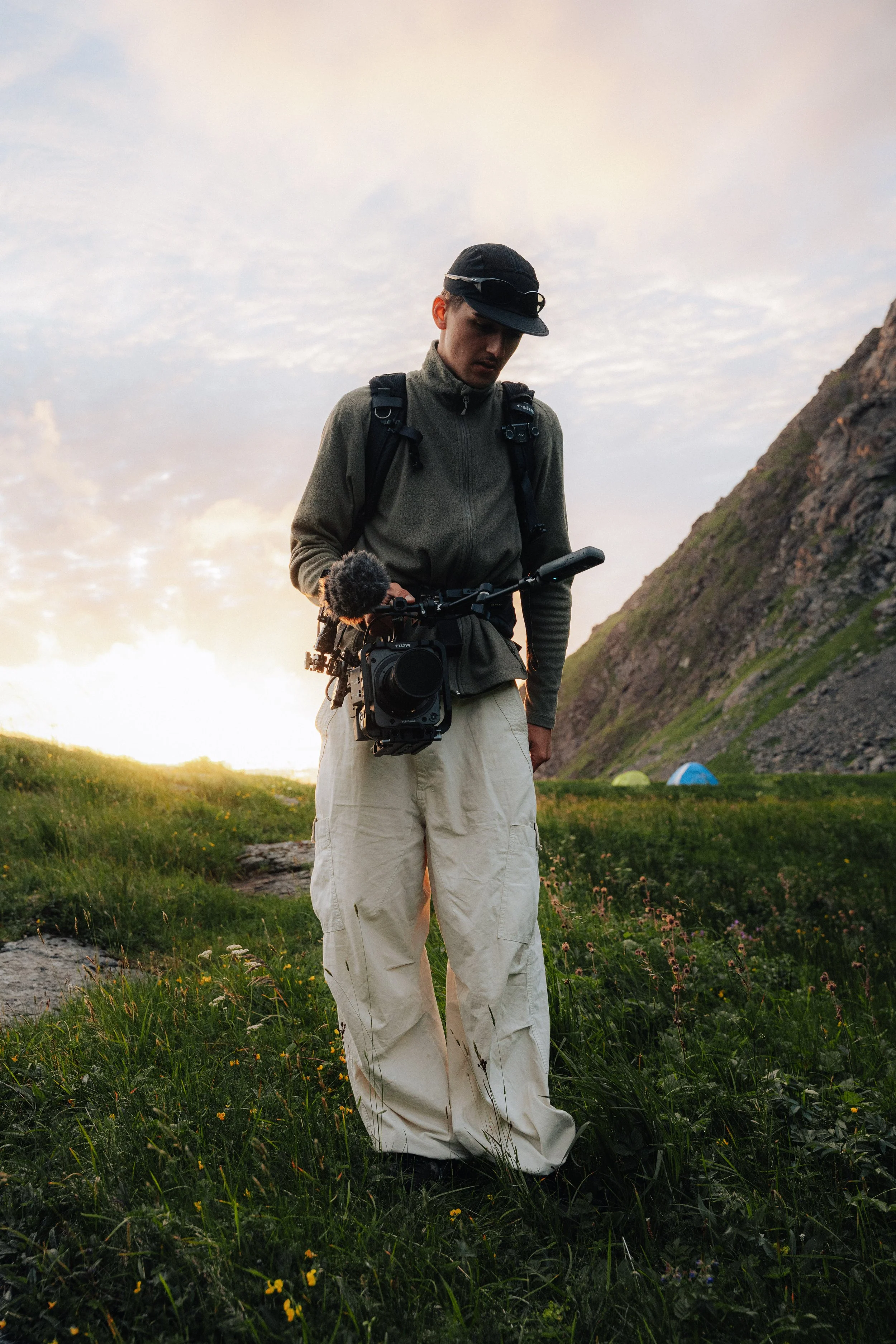 A person with a camera is standing outdoors in an open grassy field with mountains in the background during sunset.