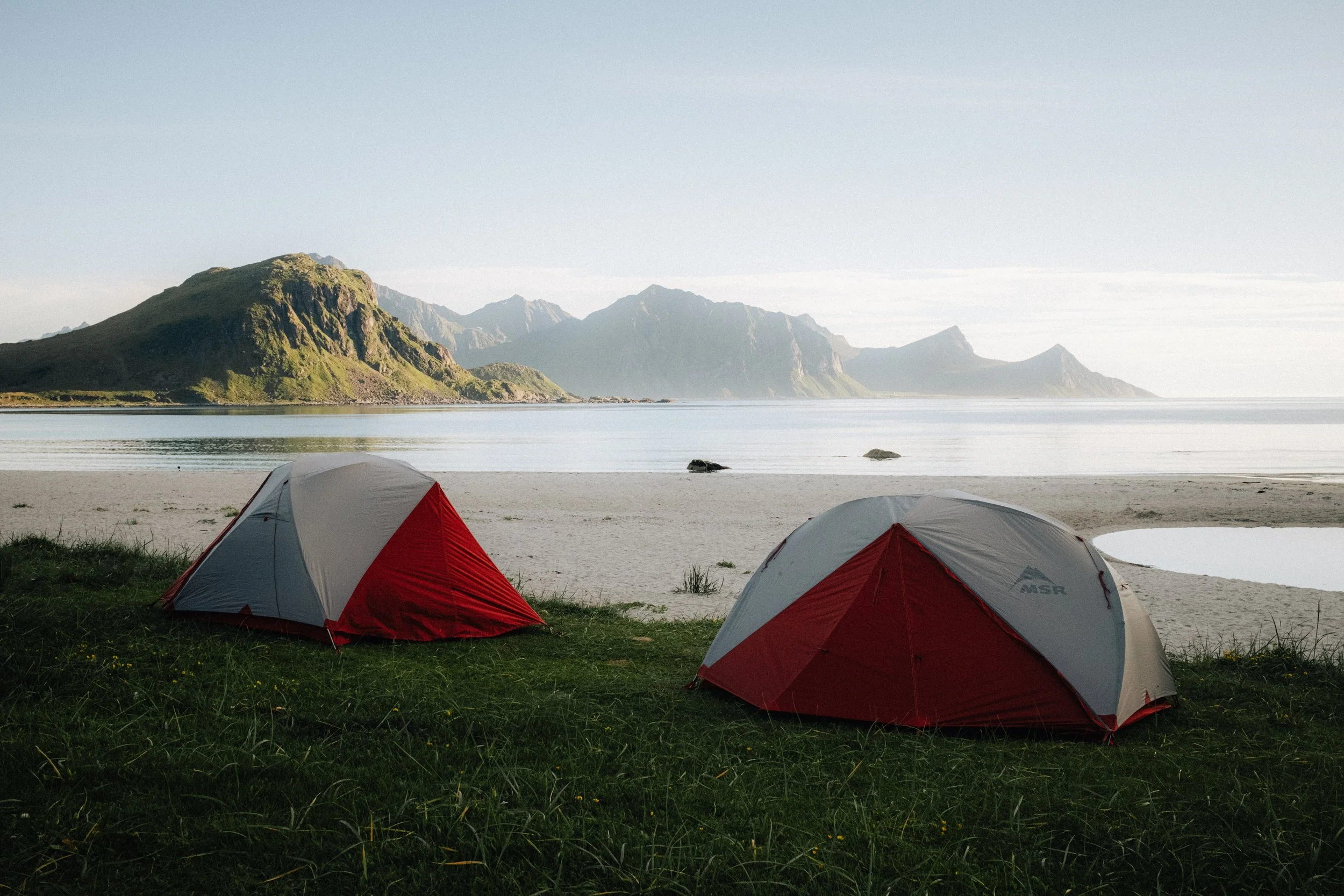 Two tents set up on green grass near a sandy beach with mountains in the background and calm water.