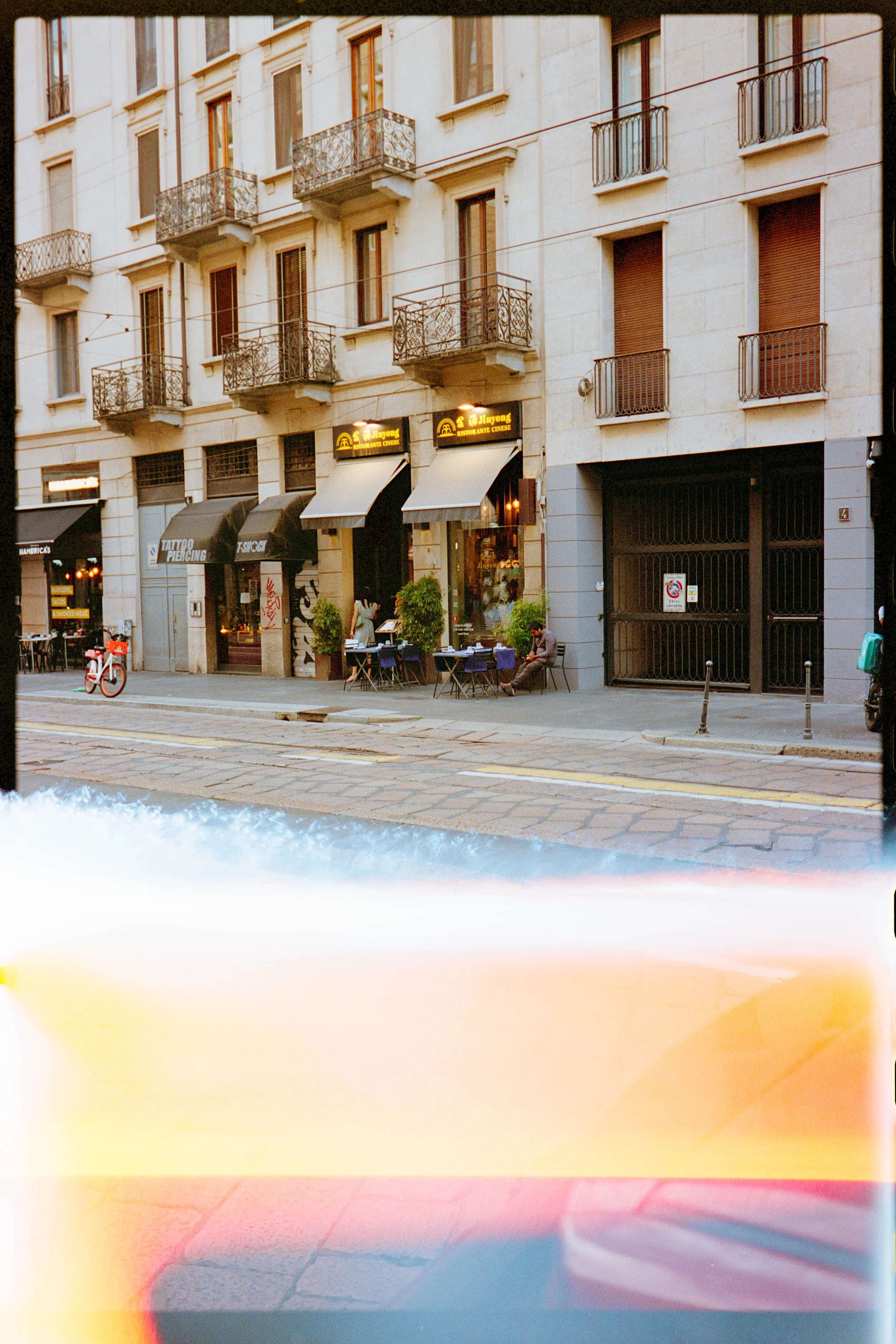 City street with a beige building, outdoor seating, a red bicycle, and a person sitting on a chair.