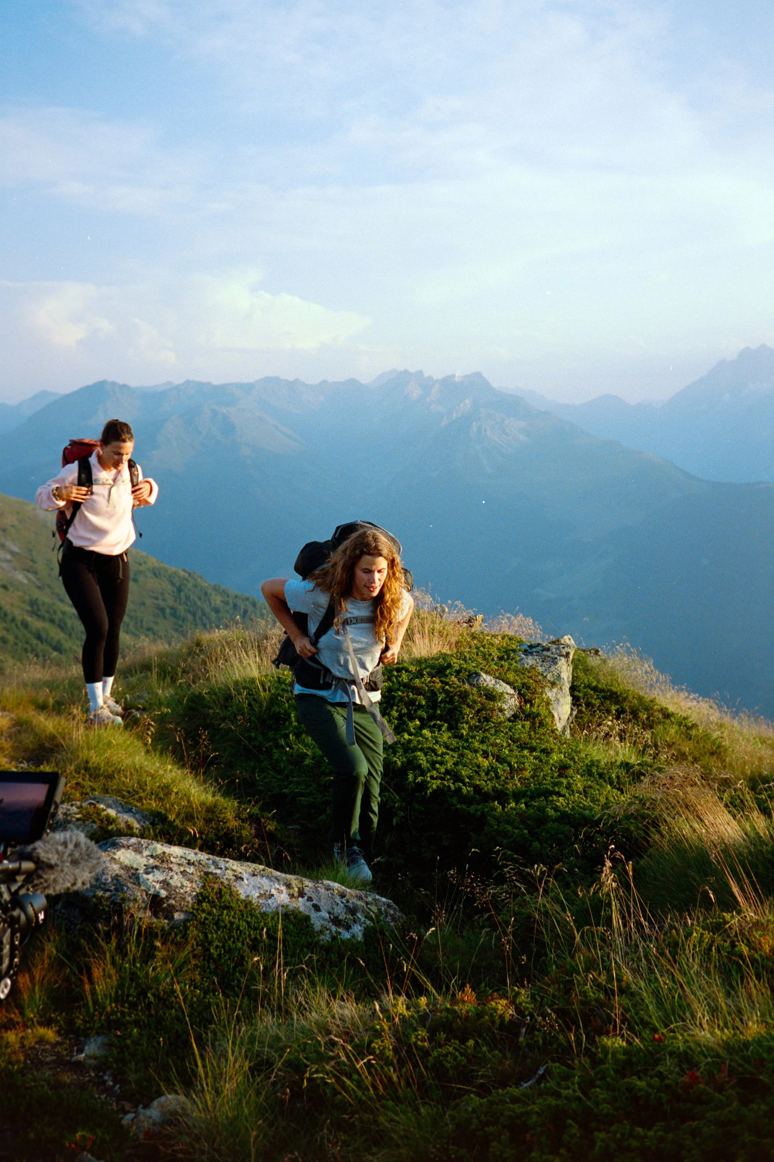 Two women hiking on a mountain trail with a mountain range in the background during daytime.