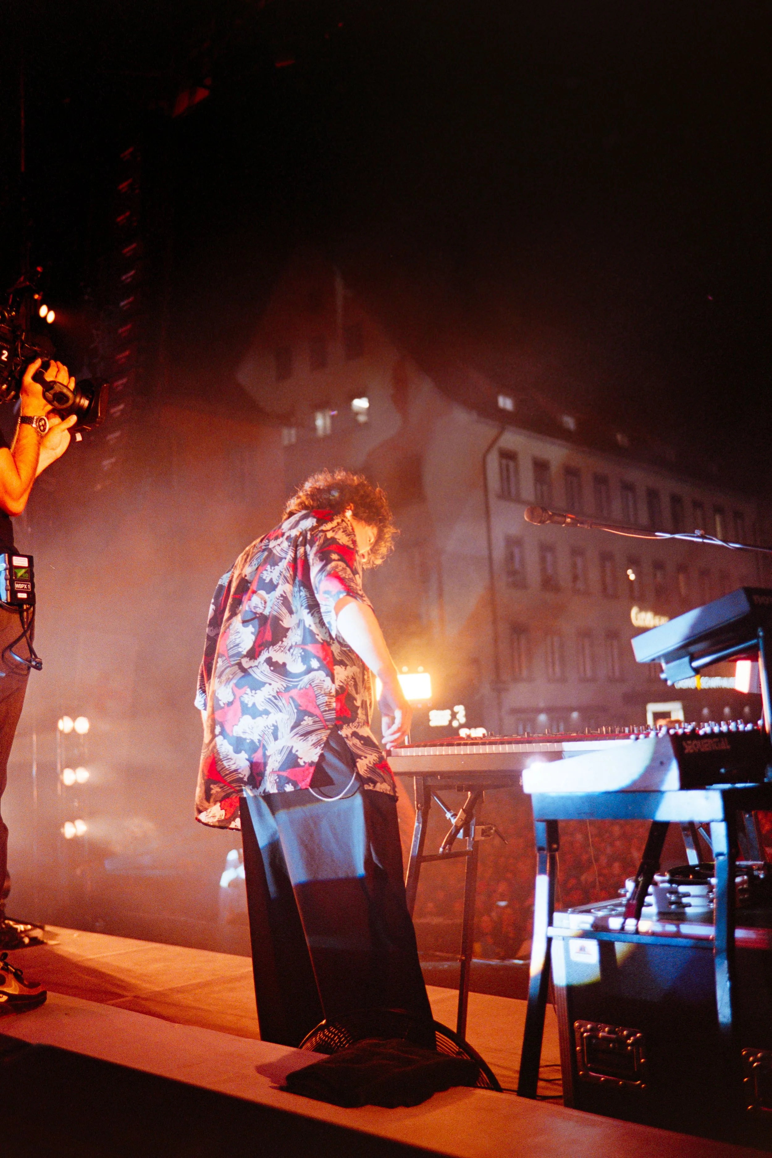 Musician playing keyboard on stage at night with city buildings in background, brightly lit, wearing a colorful patterned shirt.